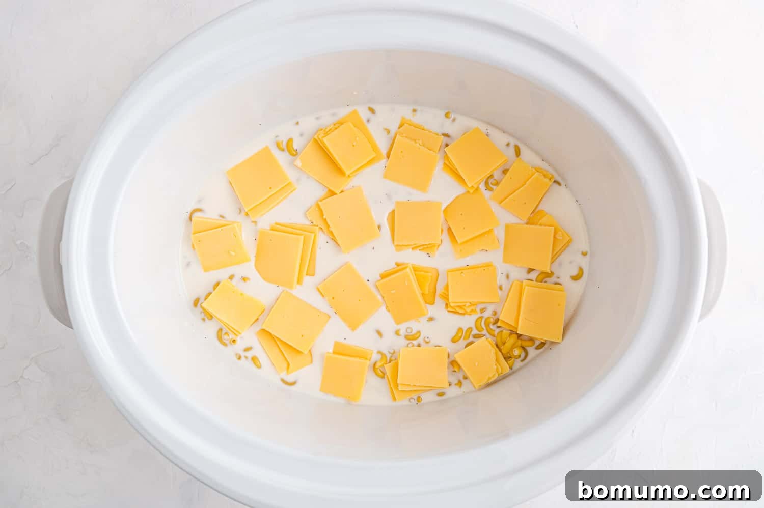 Small cubes of American cheese are carefully layered over the uncooked macaroni and liquid ingredients in a slow cooker, preparing for the next stage of cooking.