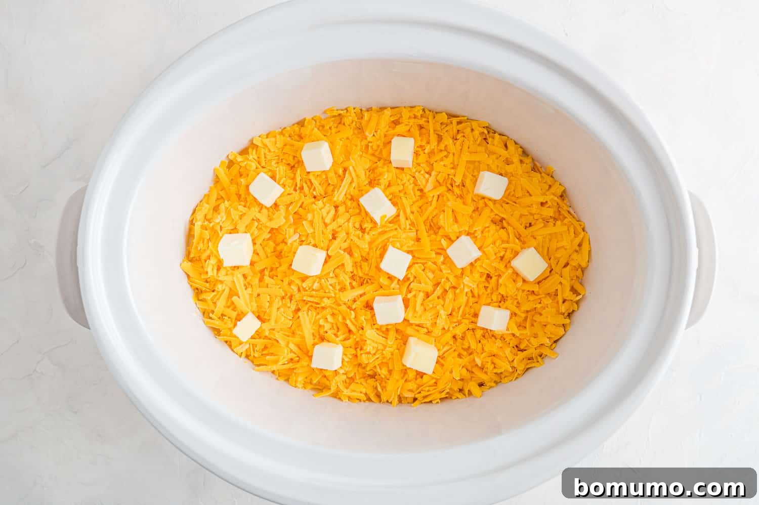 Small cubes of butter and a layer of shredded cheddar cheese are positioned on top of the macaroni and milk mixture within a slow cooker, ready for the cooking process to begin.