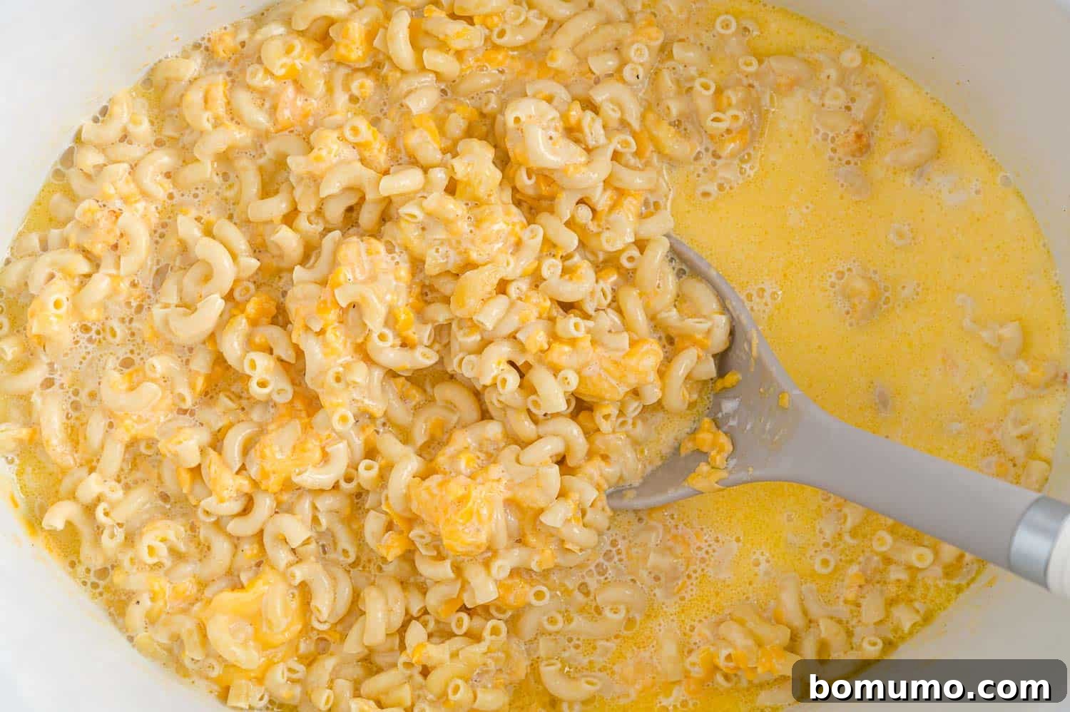 A large serving spoon stirring the mac and cheese in the slow cooker after the initial cooking period, revealing a lusciously creamy and cheesy texture forming.