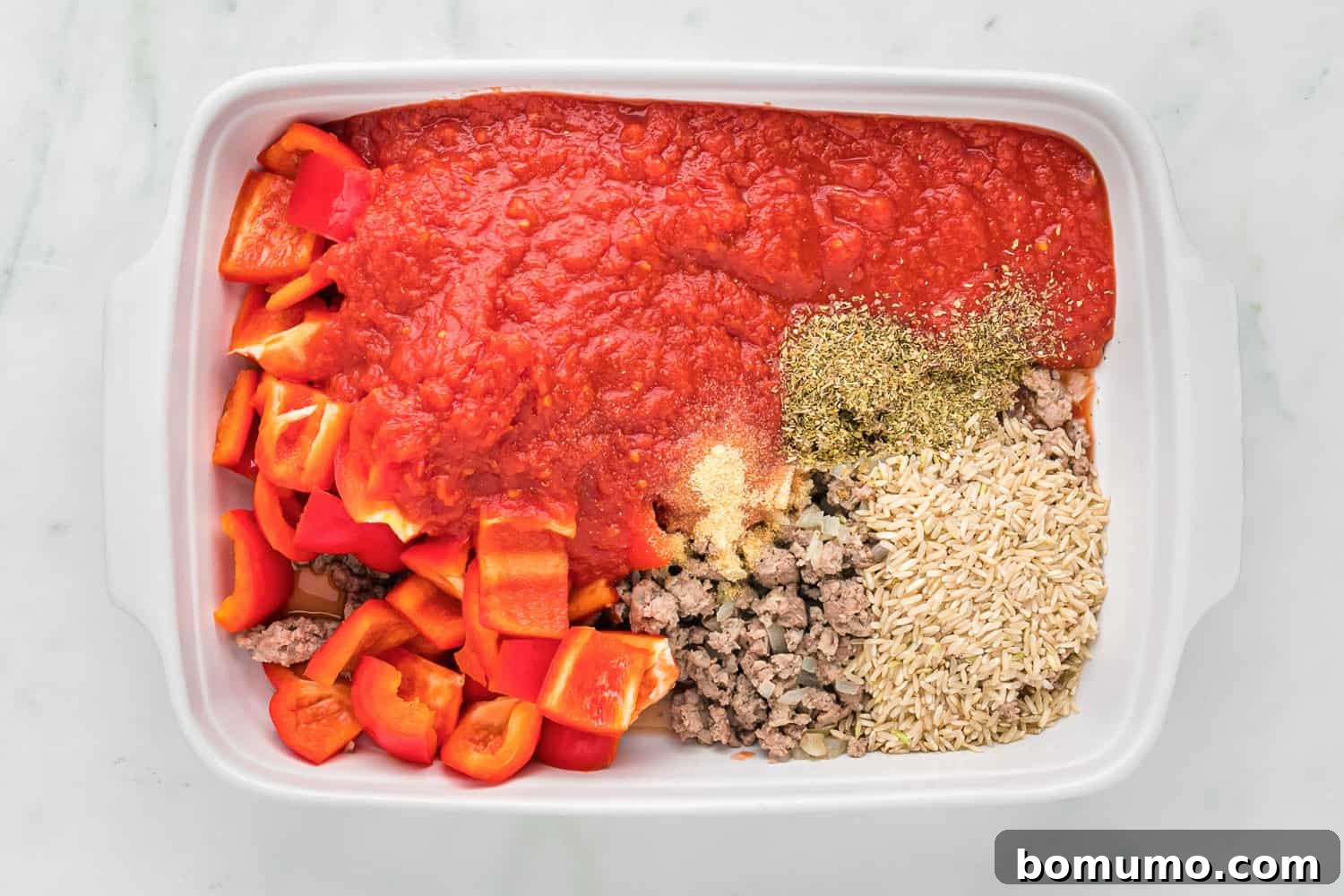 All raw ingredients including chopped bell peppers, crushed tomatoes, rice, and spices neatly arranged in a baking dish, ready for mixing.