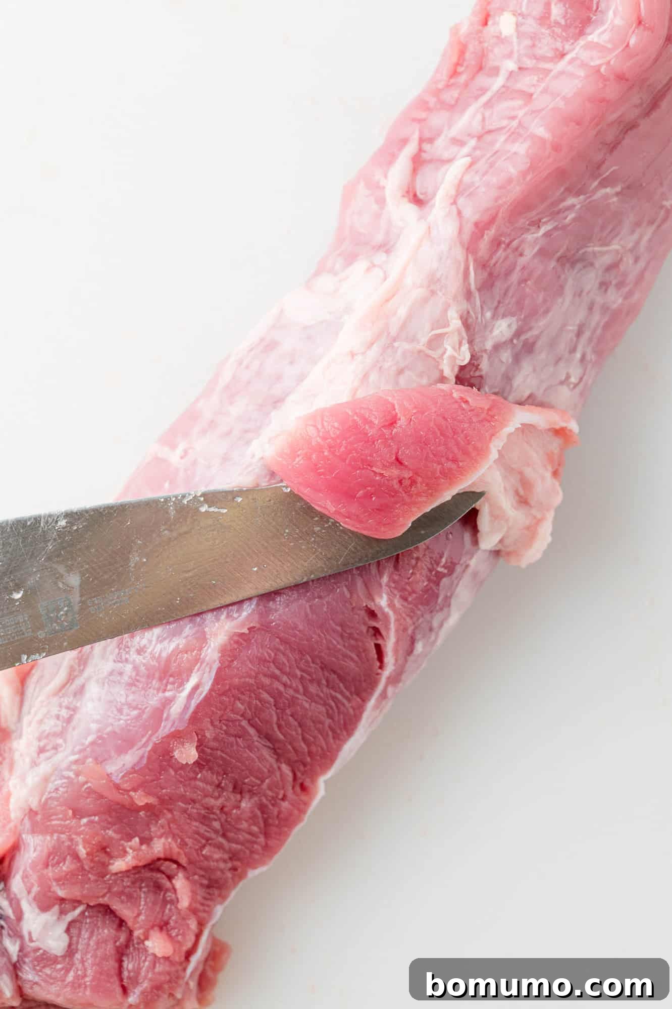 A sharp knife precisely cutting off the tough silver skin from a pork tenderloin.