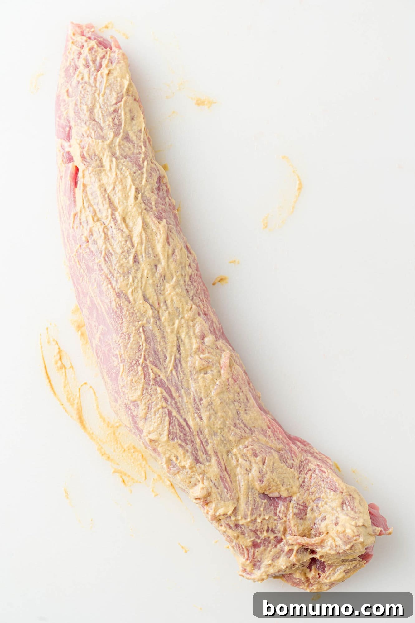 Pork tenderloin thoroughly rubbed with a vibrant layer of Dijon mustard, preparing it for the spice blend.