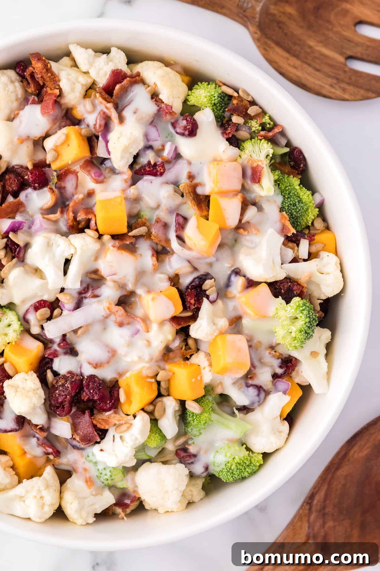 Overhead view of a finished broccoli cauliflower salad in a bowl, alongside elegant wooden salad scoops.