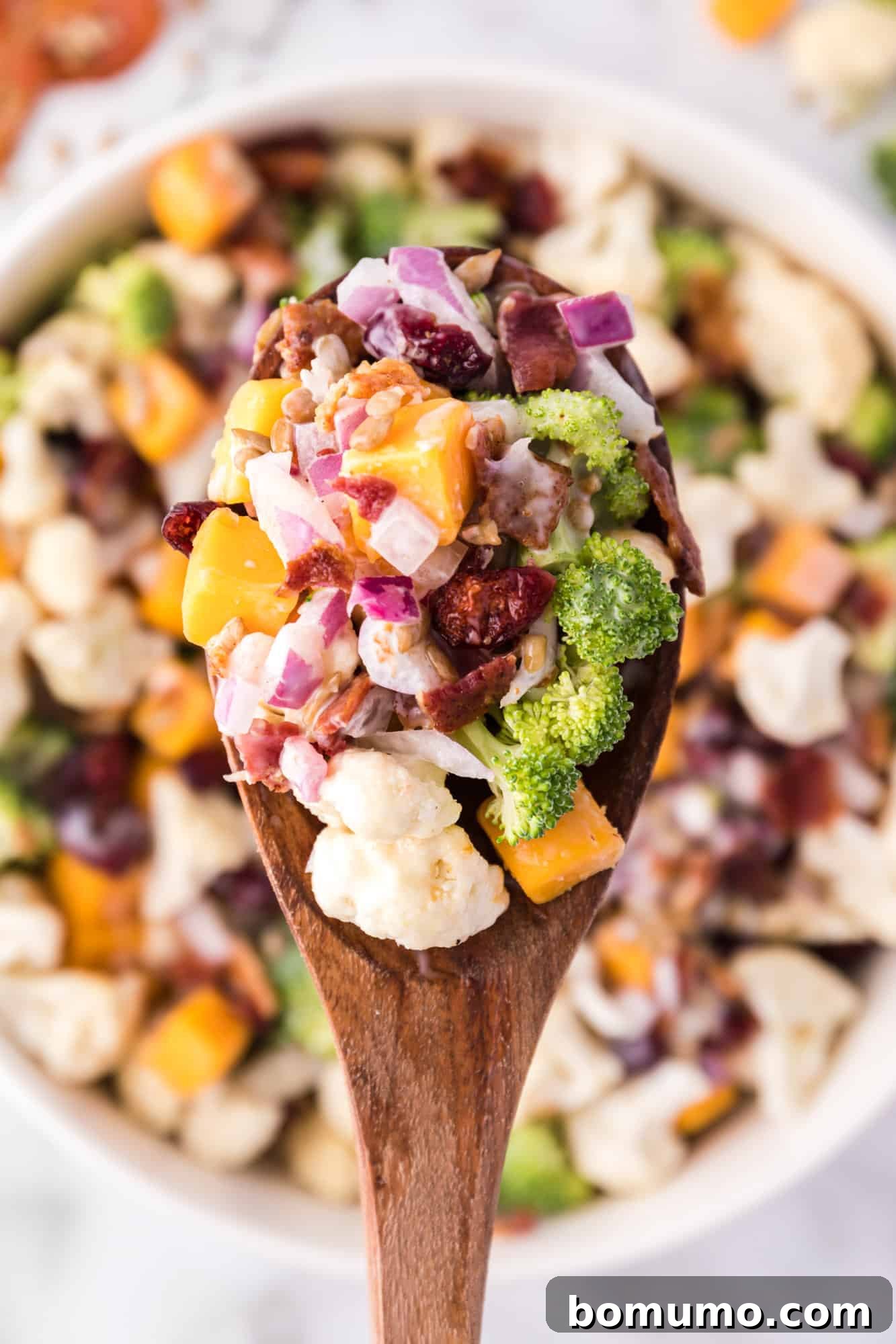 Close up overhead view of a large spoonful of creamy broccoli cauliflower salad held over a bowl, highlighting its texture and ingredients.