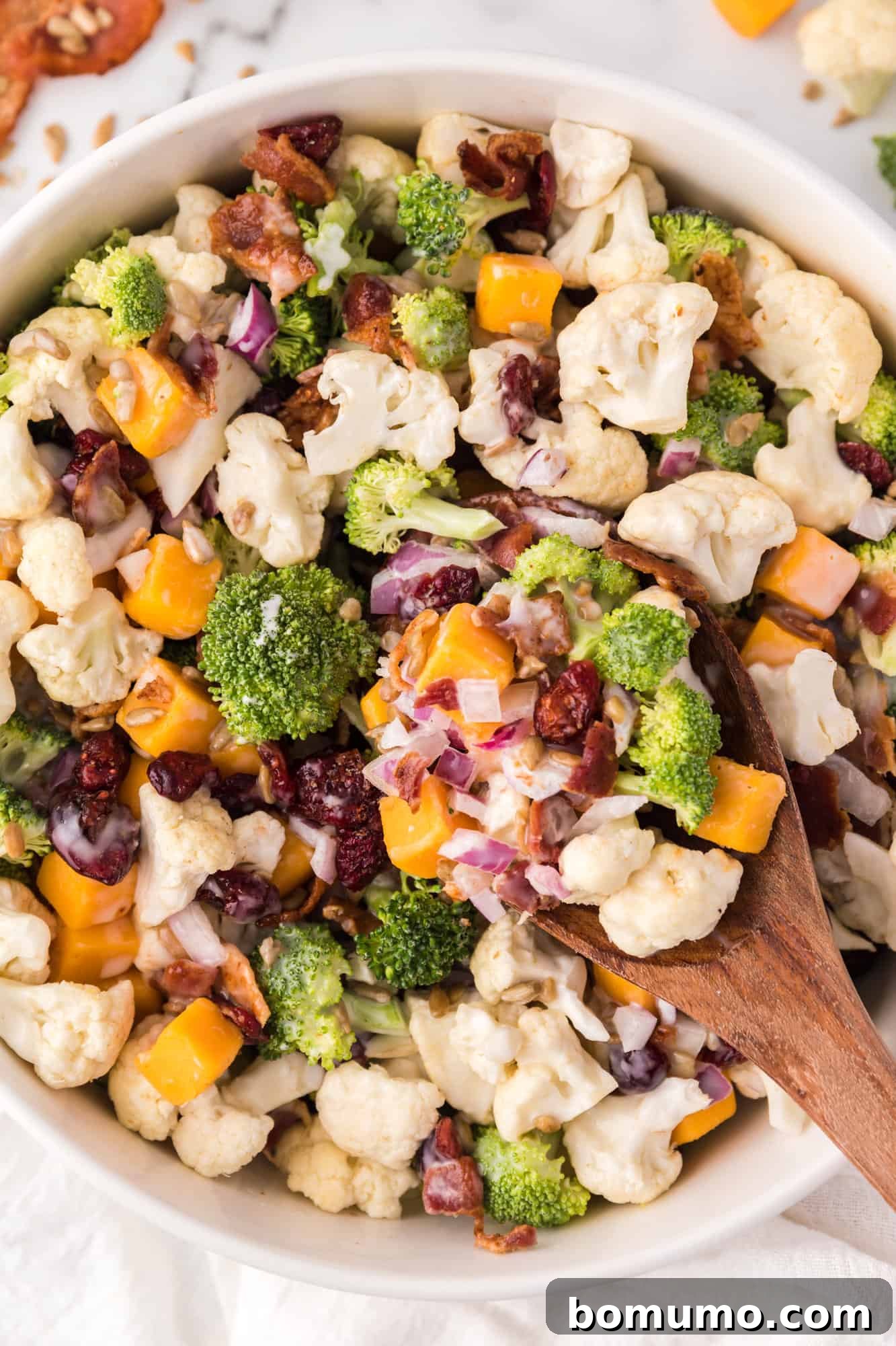 Overhead view of a vibrant broccoli cauliflower salad in a white bowl, served with a wooden spoon.