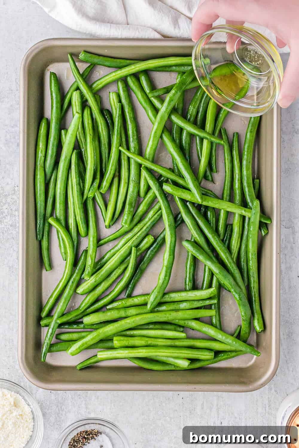 Person pouring olive oil over fresh green beans arranged on a baking tray, with small bowls of ingredients like pepper and cheese nearby.