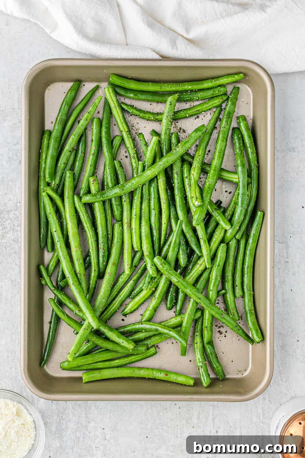 A baking tray filled with raw green beans seasoned with pepper. A white cloth is partially visible on the top edge.