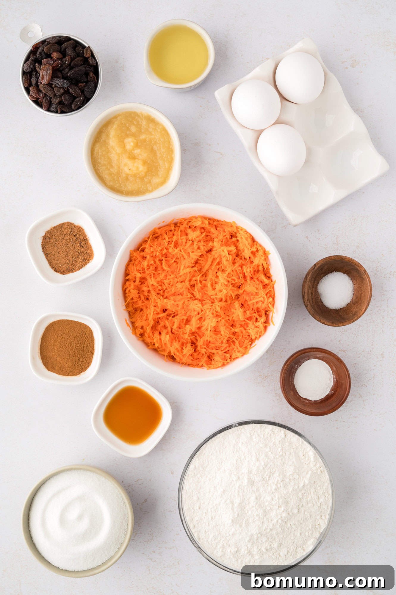 Overhead view of all fresh ingredients neatly laid out for making carrot cake muffins, including flour, eggs, carrots, and spices.