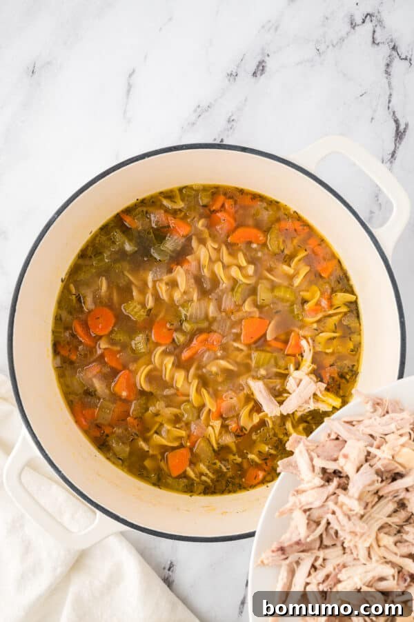 Shredded chicken being gently poured back into the simmering pot of chicken noodle soup, ready to combine with noodles and broth.