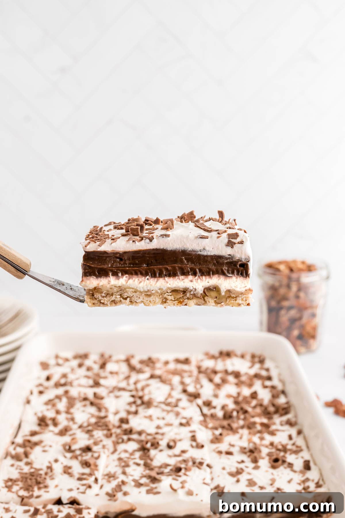 A perfectly sliced piece of Chocolate Delight dessert, featuring visible layers of pecan crust, cream cheese, chocolate pudding, and whipped topping, held on a spatula over the rest of the dessert.