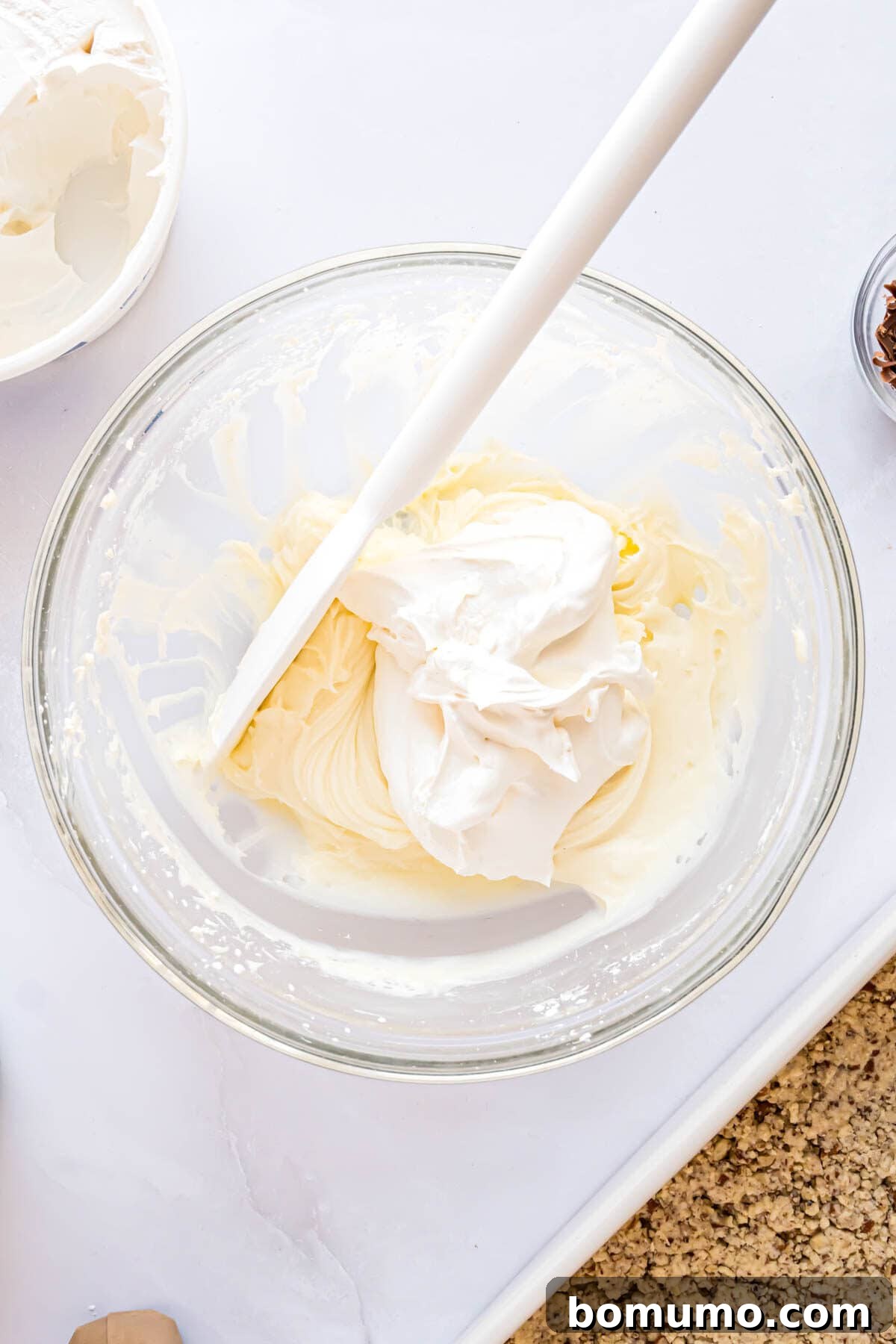Thawed whipped cream being gently folded into the cream cheese mixture by hand, maintaining its airy texture.