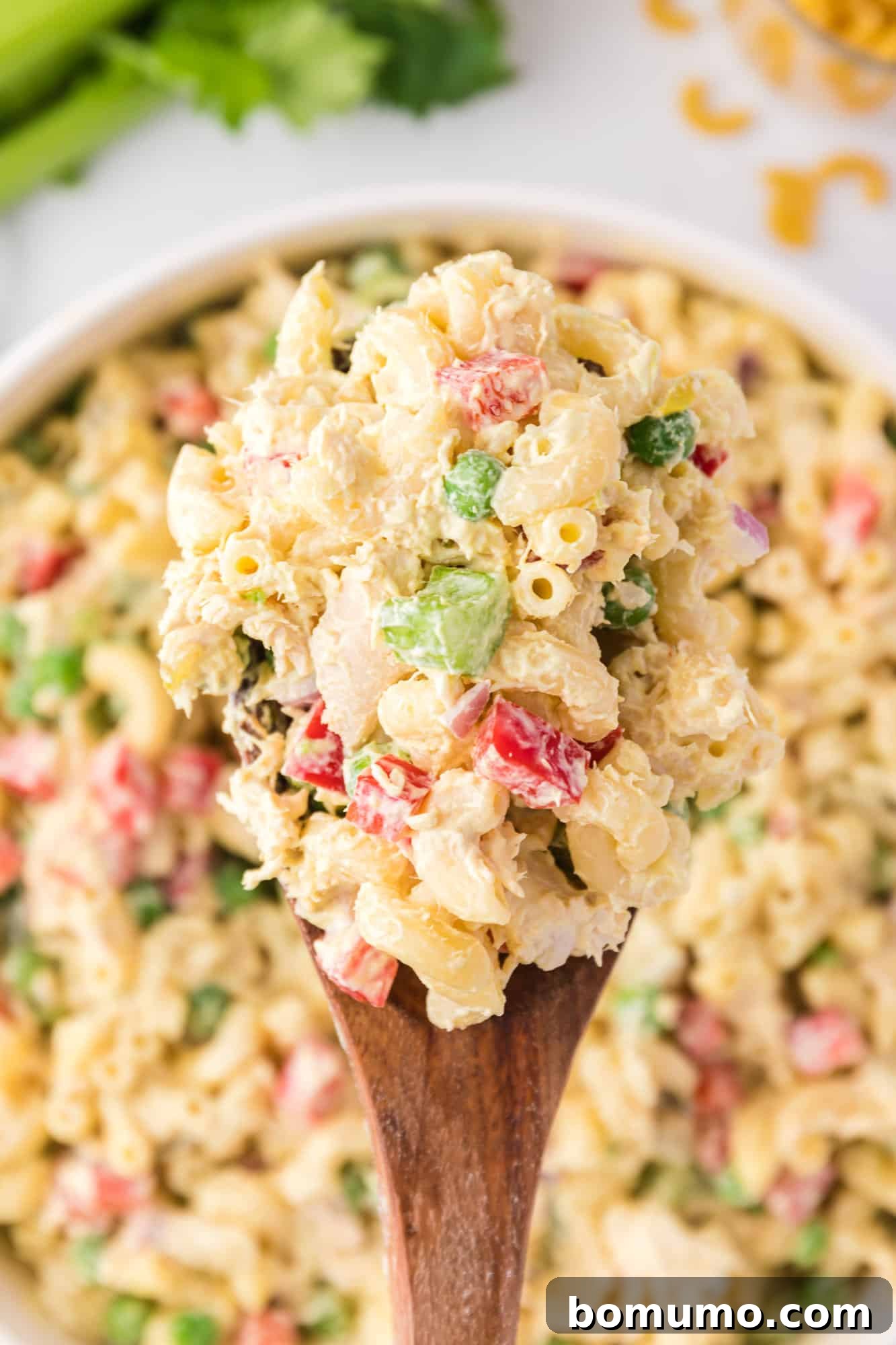 Overhead view of a spoonful of chicken macaroni salad held over a large bowl, showcasing its texture.
