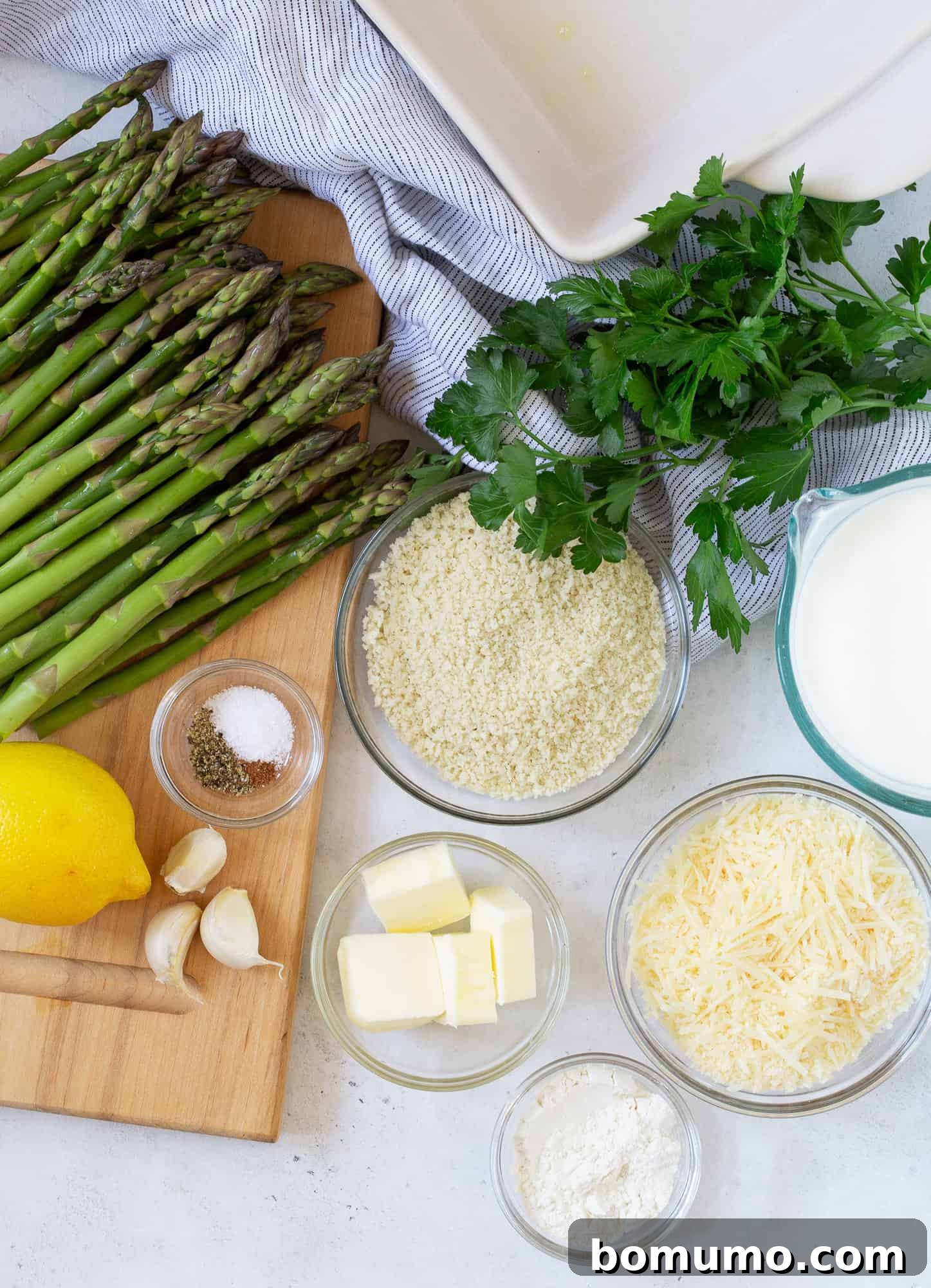 Creamy Asparagus Bake 3 Overhead view of fresh ingredients arranged for making the asparagus casserole, including vibrant green asparagus spears, a lemon, a bunch of parsley, garlic, and cheese.
