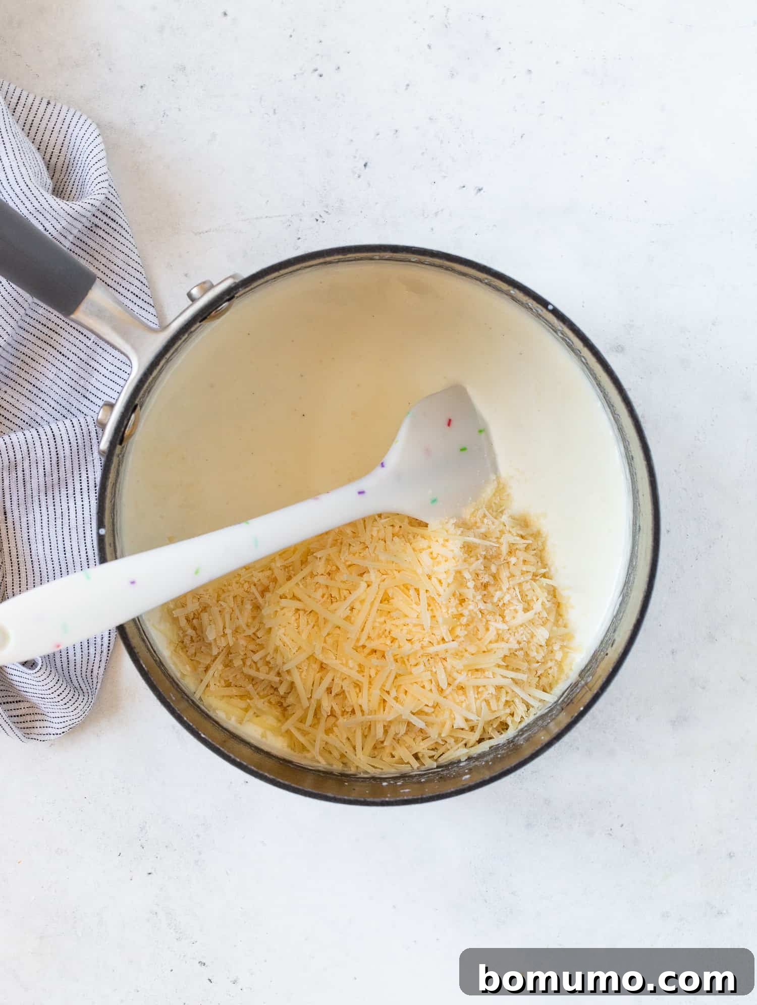 Creamy Asparagus Bake 10 Shredded Parmesan cheese being stirred into the white sauce in a saucepan.