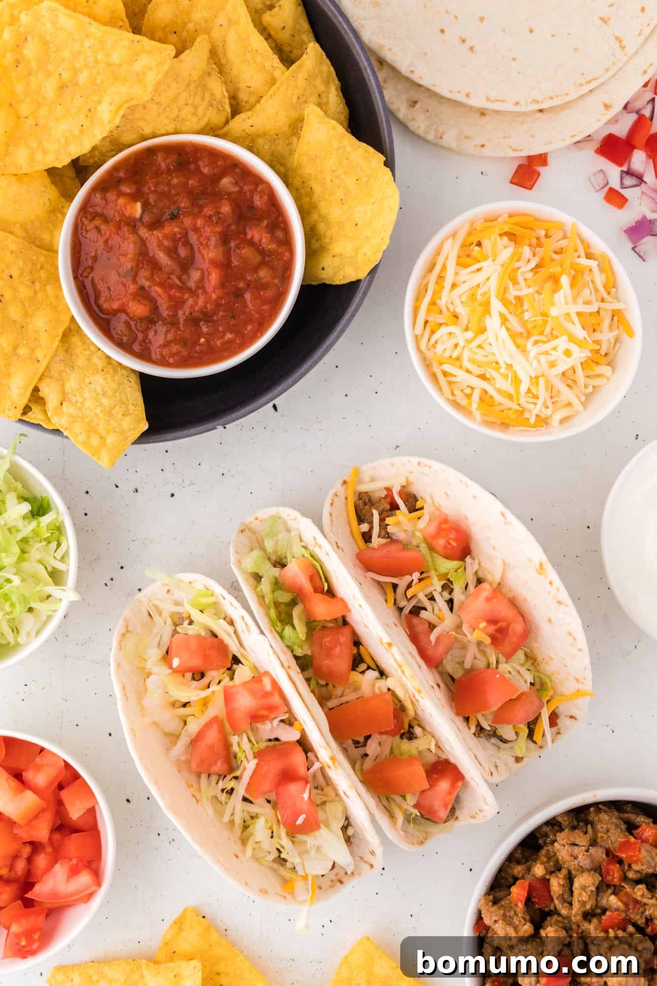 Overhead view of a taco night spread including three assembled turkey tacos, a plate of tortilla chips and salsa, and bowls of various taco toppings.