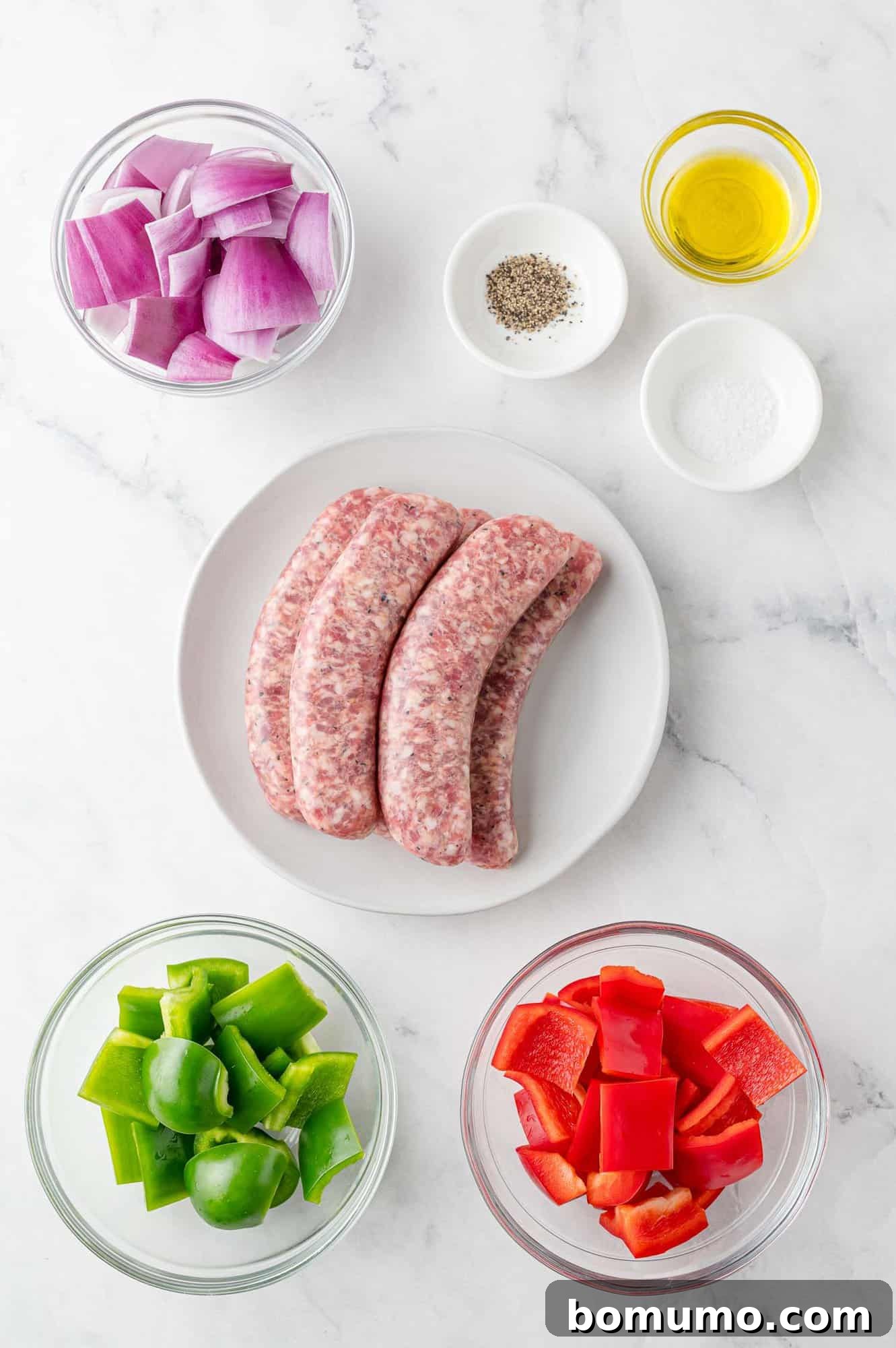 Overhead view of fresh ingredients laid out: raw Italian sausage links, colorful bell peppers, a red onion, and olive oil with salt and pepper, ready for preparation.