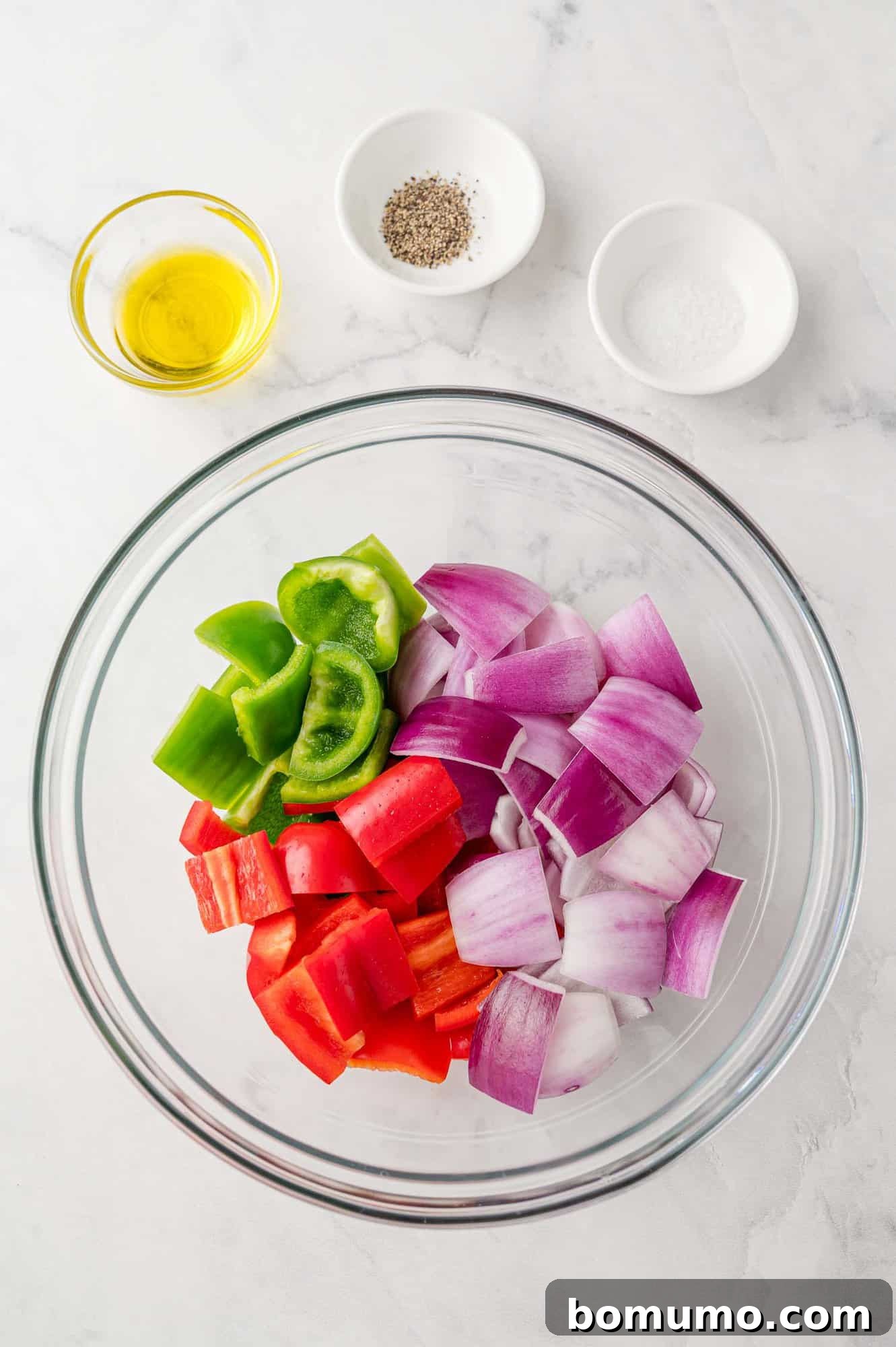 Freshly chopped bell peppers and red onions in a mixing bowl, alongside olive oil, salt, and pepper, awaiting seasoning.
