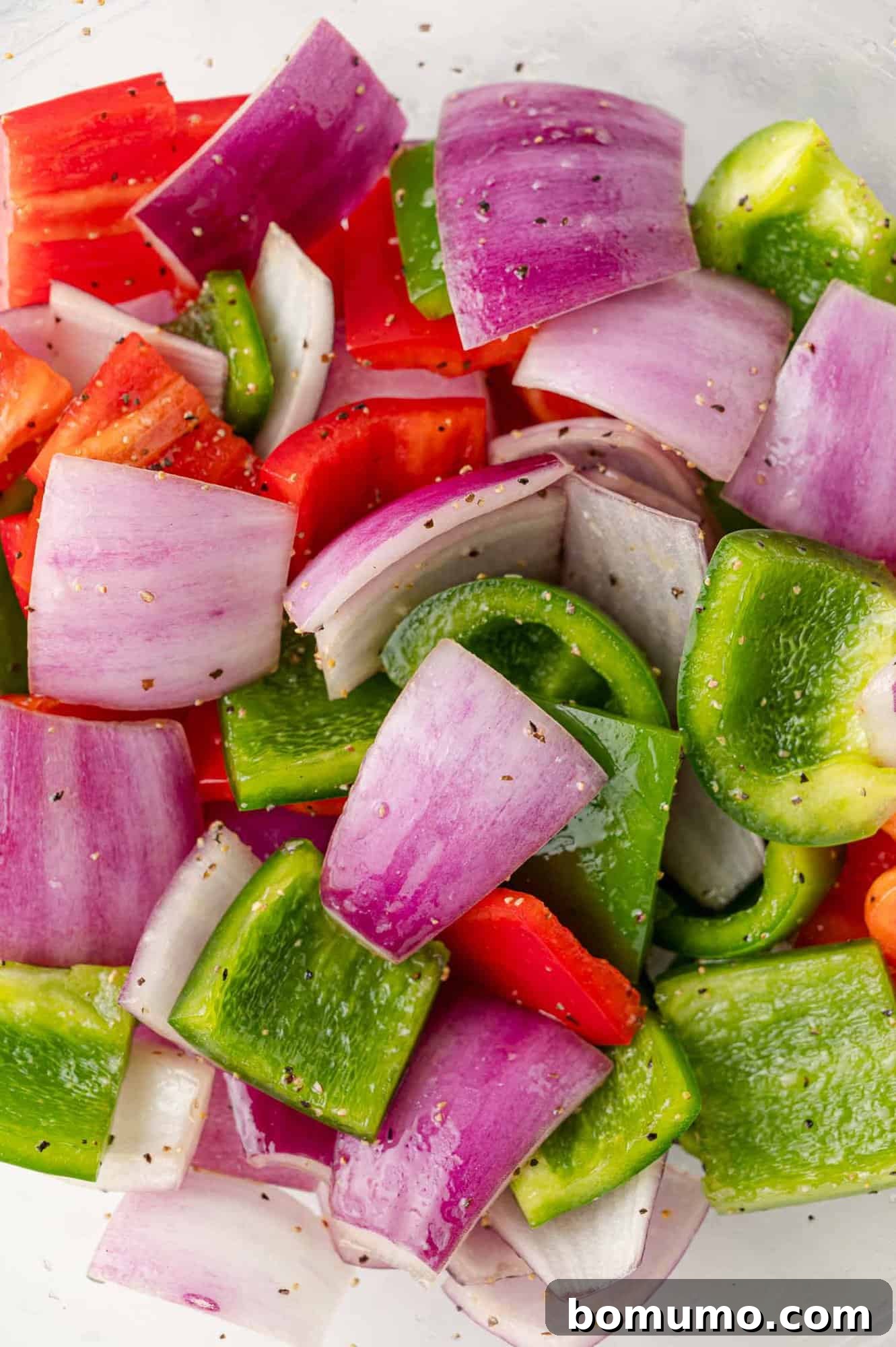 Vibrant bell peppers and red onions coated with olive oil and seasonings, perfectly mixed in a bowl and ready to be spread on the baking sheet.