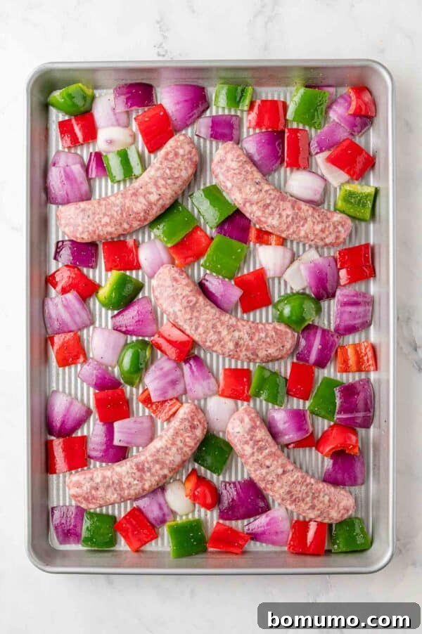 Raw Italian sausage links laid out on a sheet pan surrounded by colorful bell peppers and red onions, ready for the oven.