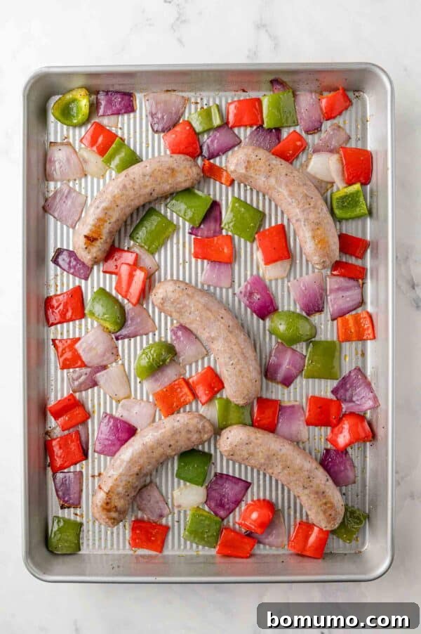 Partially cooked Italian sausage with peppers and onions on a sheet pan, showing initial browning after 15 minutes in the oven.