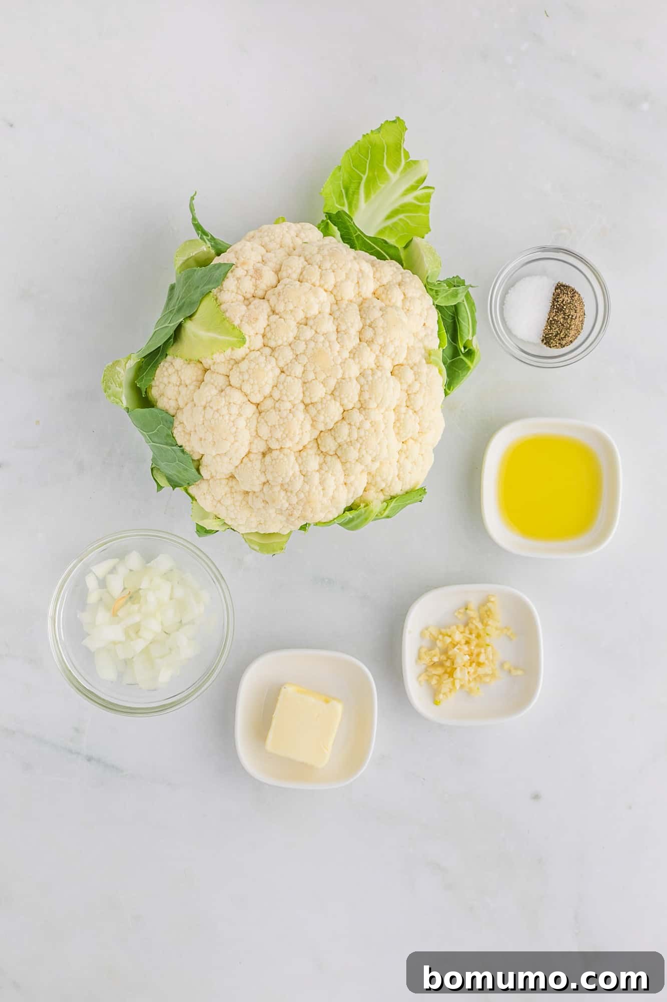 Ingredients laid out on a kitchen counter for making cauliflower rice, including a whole head of cauliflower, garlic, onion, butter, and olive oil.