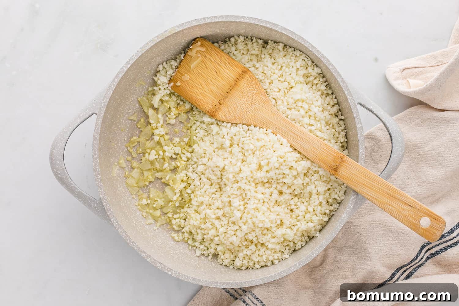 Raw riced cauliflower cooking in a skillet with sautéed onions and garlic, preparing to become a flavorful side dish.