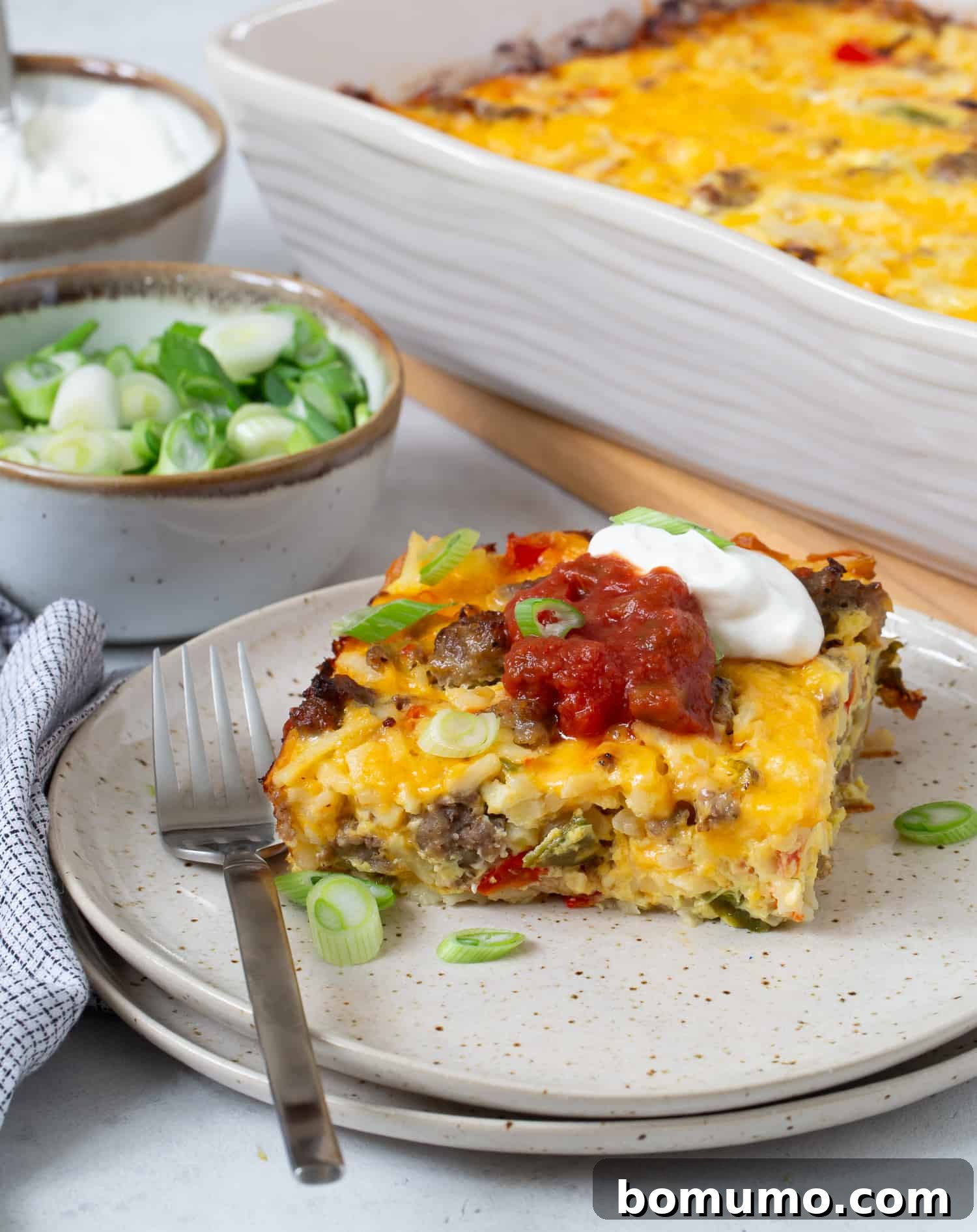 Slice of hash brown breakfast casserole topped with salsa and sour cream, remaining casserole in background.