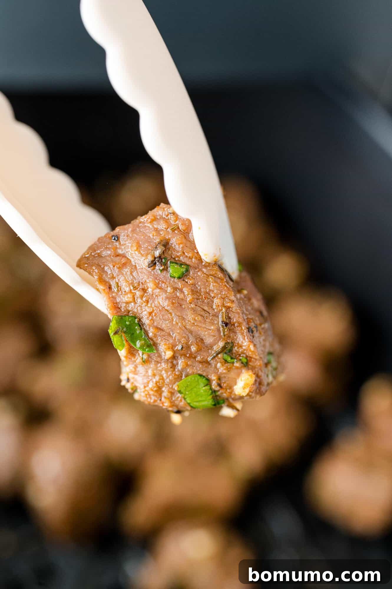 A single air fryer steak bite, held by tongs, showcasing its perfect doneness and inviting texture, lifted above a pile of more steak bites.
