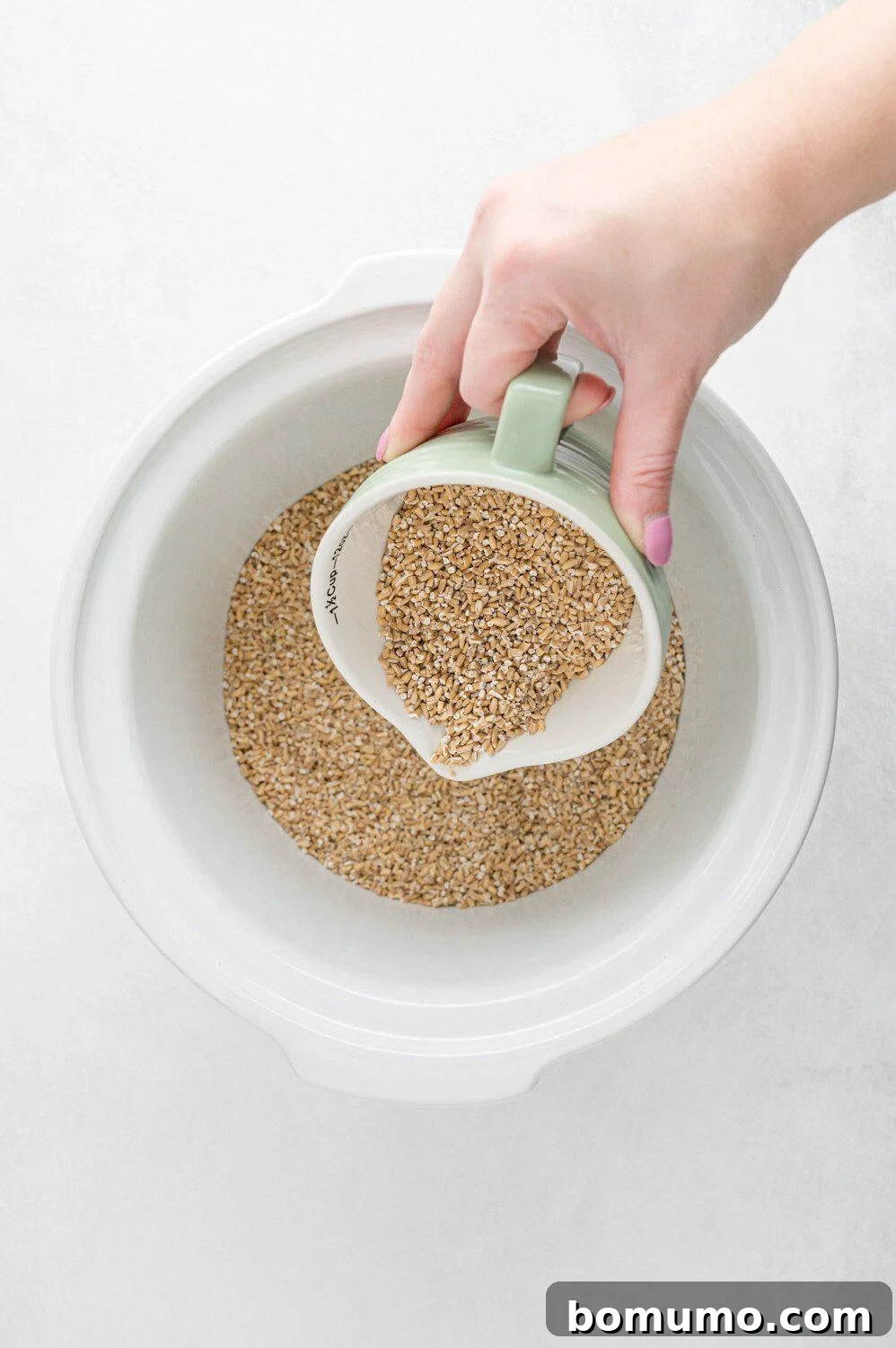 A hand carefully pours steel cut oats from a light green measuring cup into a pristine white slow cooker bowl.