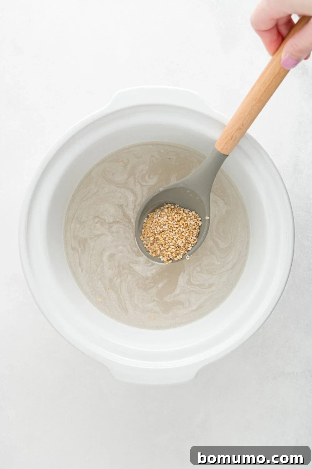 A hand holds a clear measuring cup, pouring fresh water into a white bowl filled with uncooked oats, preparing them for a wholesome meal.
