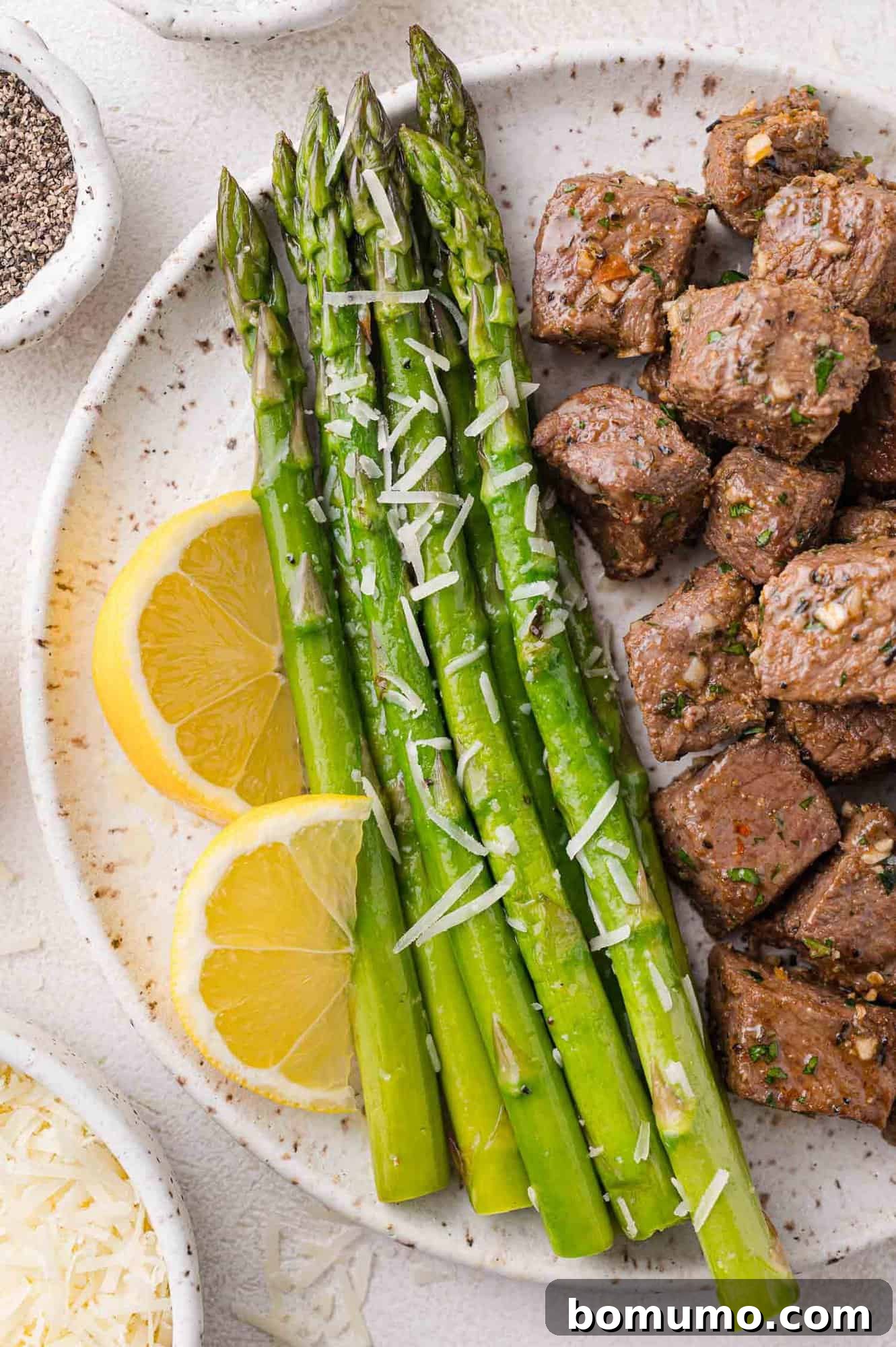 Sautéed asparagus spears on a plate, served with garlic butter steak bites and lemon slices.