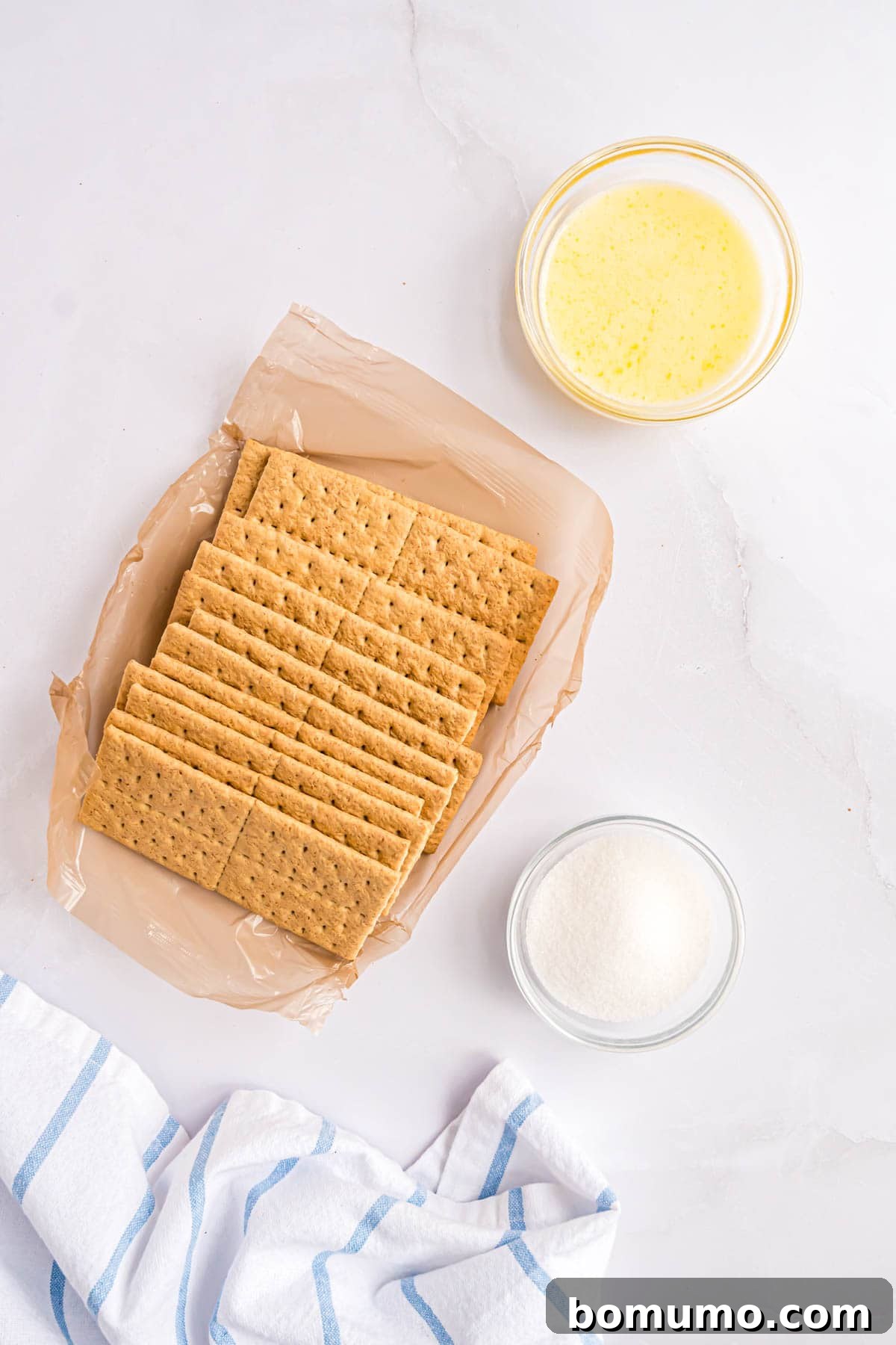 Overhead shot of three key ingredients for graham cracker crust: whole graham crackers, a bowl of granulated sugar, and a bowl of melted butter.
