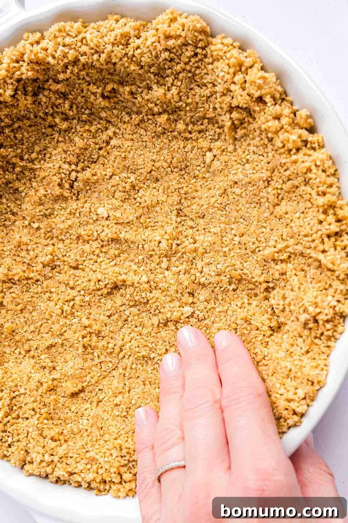 A hand gently pressing the graham cracker crumb mixture into the bottom of a pie plate.