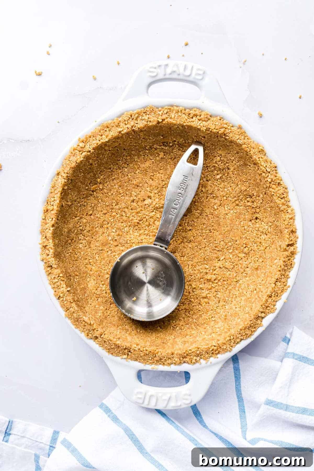 The bottom of a measuring cup being used to firmly press graham cracker crust into a pie plate, ensuring even compaction.