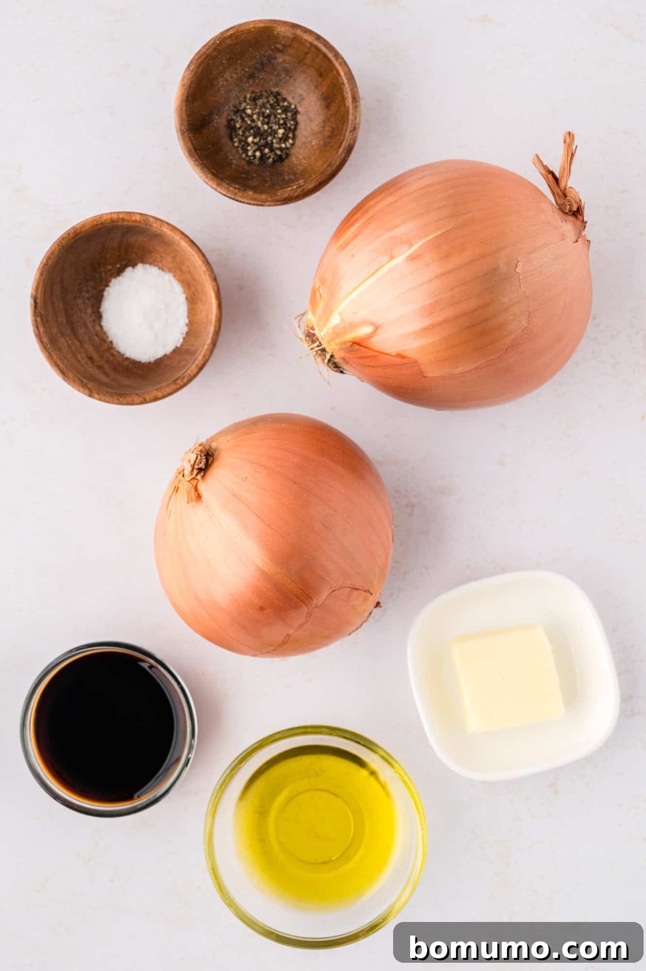 Fresh ingredients laid out on a wooden cutting board, including two large, unpeeled onions, a bottle of olive oil, a stick of butter, and a pepper grinder, showcasing readiness for cooking sautéed onions.