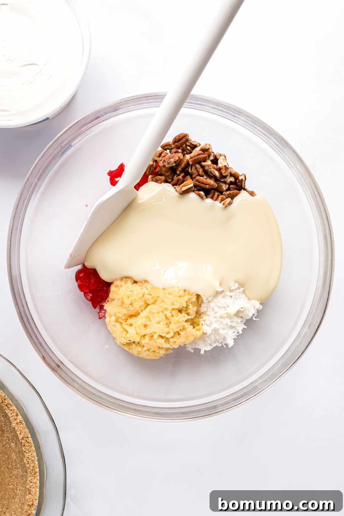 Golden Treasure Pie 5 A clear glass bowl containing all the Millionaire Pie filling ingredients, excluding the whipped cream, before they have been mixed together. The distinct layers of fruit, nuts, and condensed milk are visible.