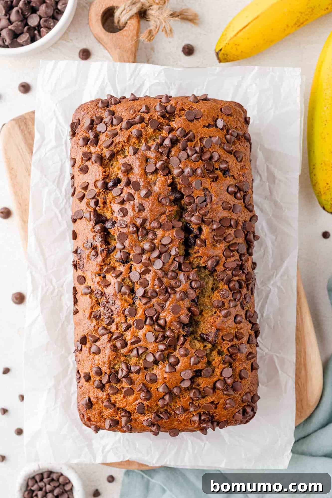 A perfectly baked loaf of chocolate chip banana bread, cooling on a wire rack, surrounded by fresh bananas and extra chocolate chips.