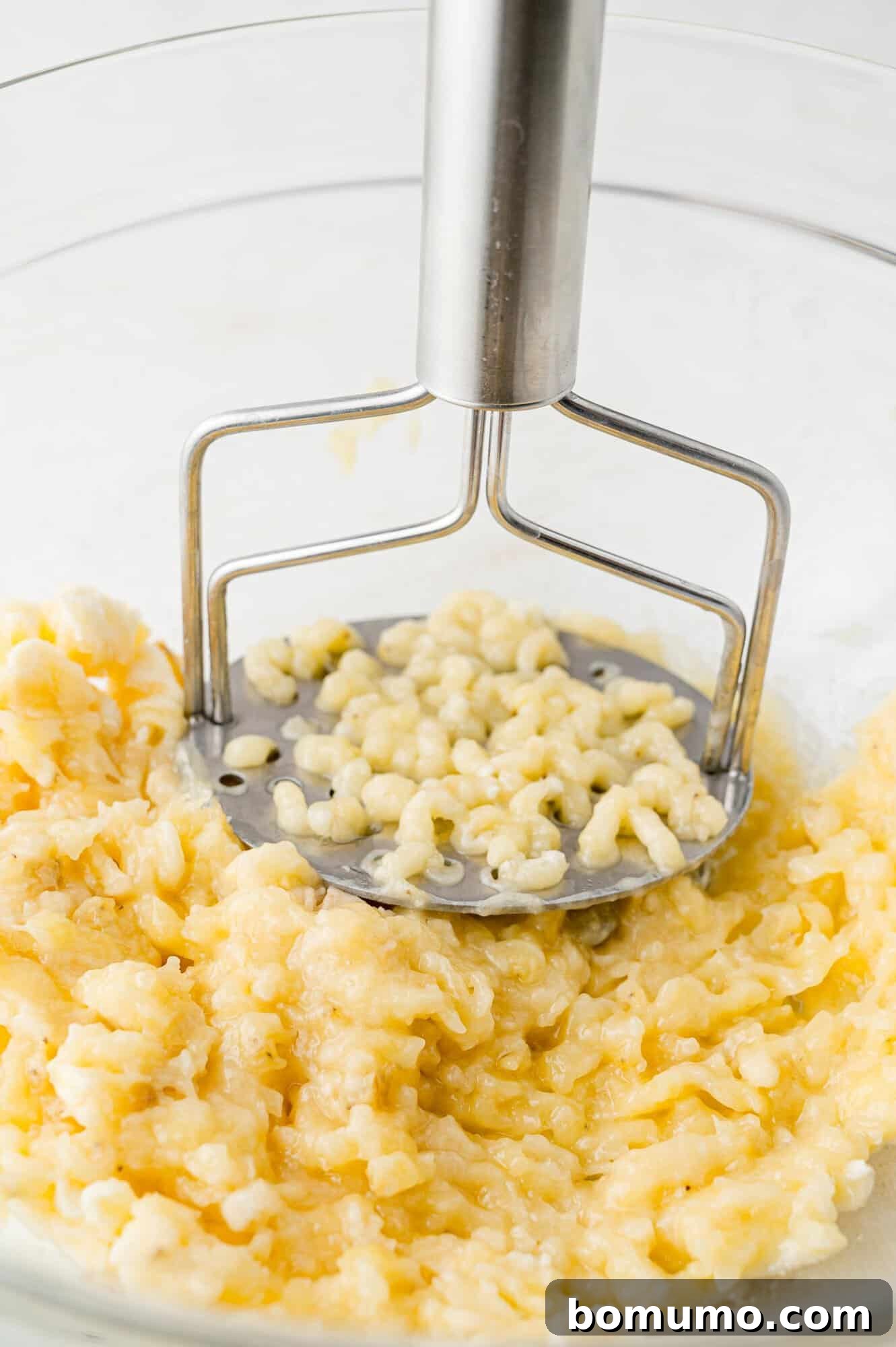 Ripe bananas being mashed with a potato masher in a large, clear glass mixing bowl.