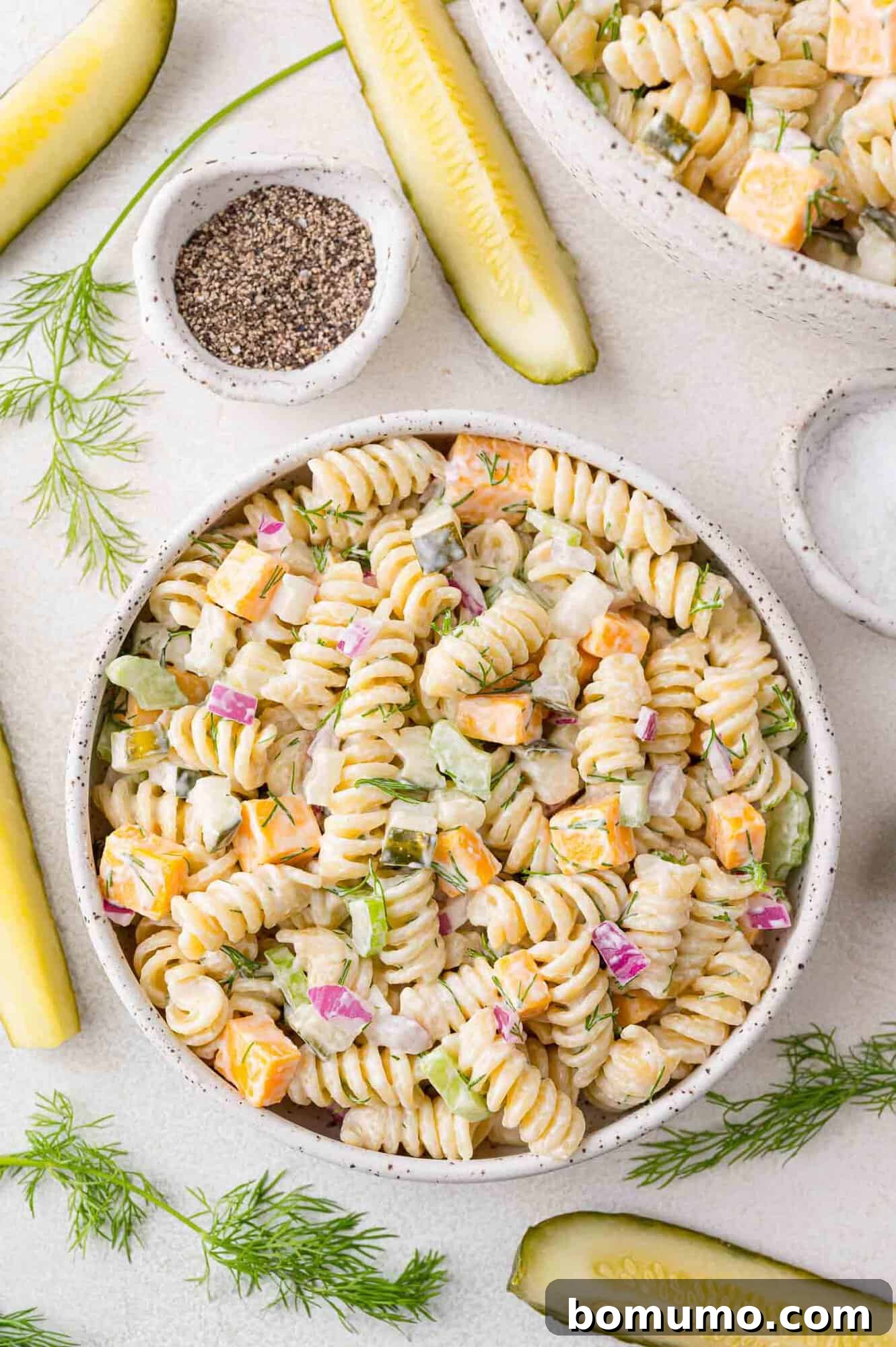 Overhead view of dill pickle pasta salad in a large bowl, with whole pickles and fresh dill sprigs arranged around it.