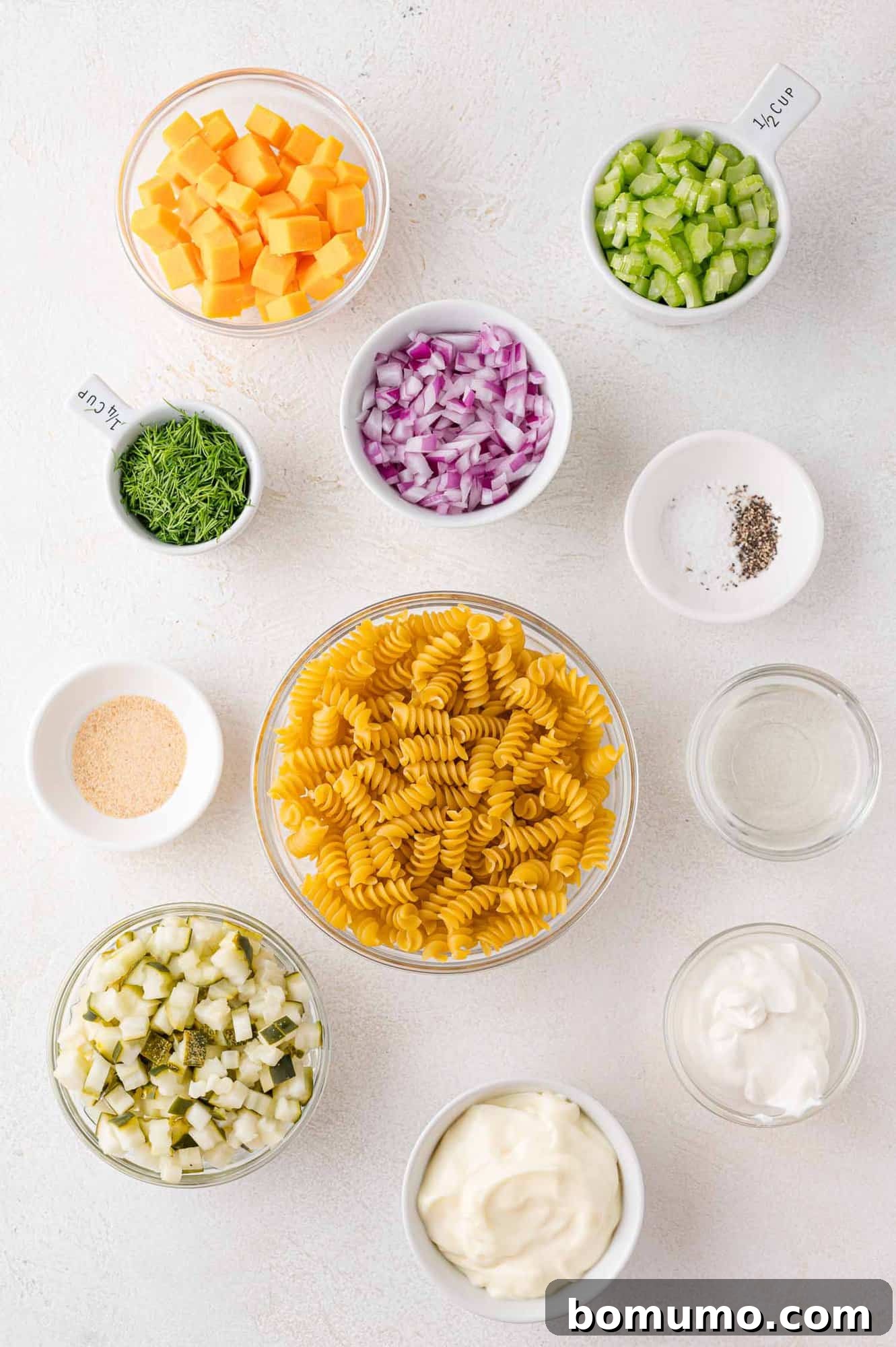Ingredients needed for Dill Pickle Pasta Salad in small separate bowls, showcasing fresh dill, diced pickles, cheese, and pasta.