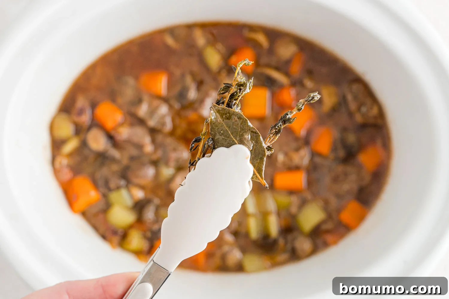 A hand carefully removing the bay leaf from the rich, simmering beef stew in a slow cooker.