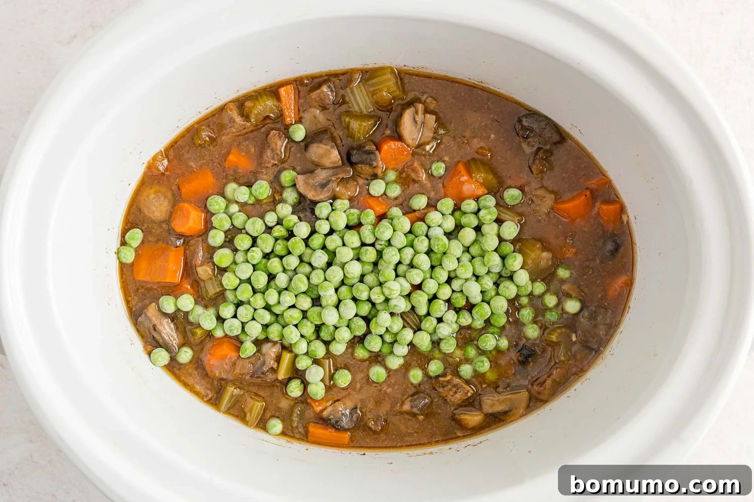 Vibrant green frozen peas being stirred into the hot beef Marsala stew in a crockpot, adding a burst of color and freshness.
