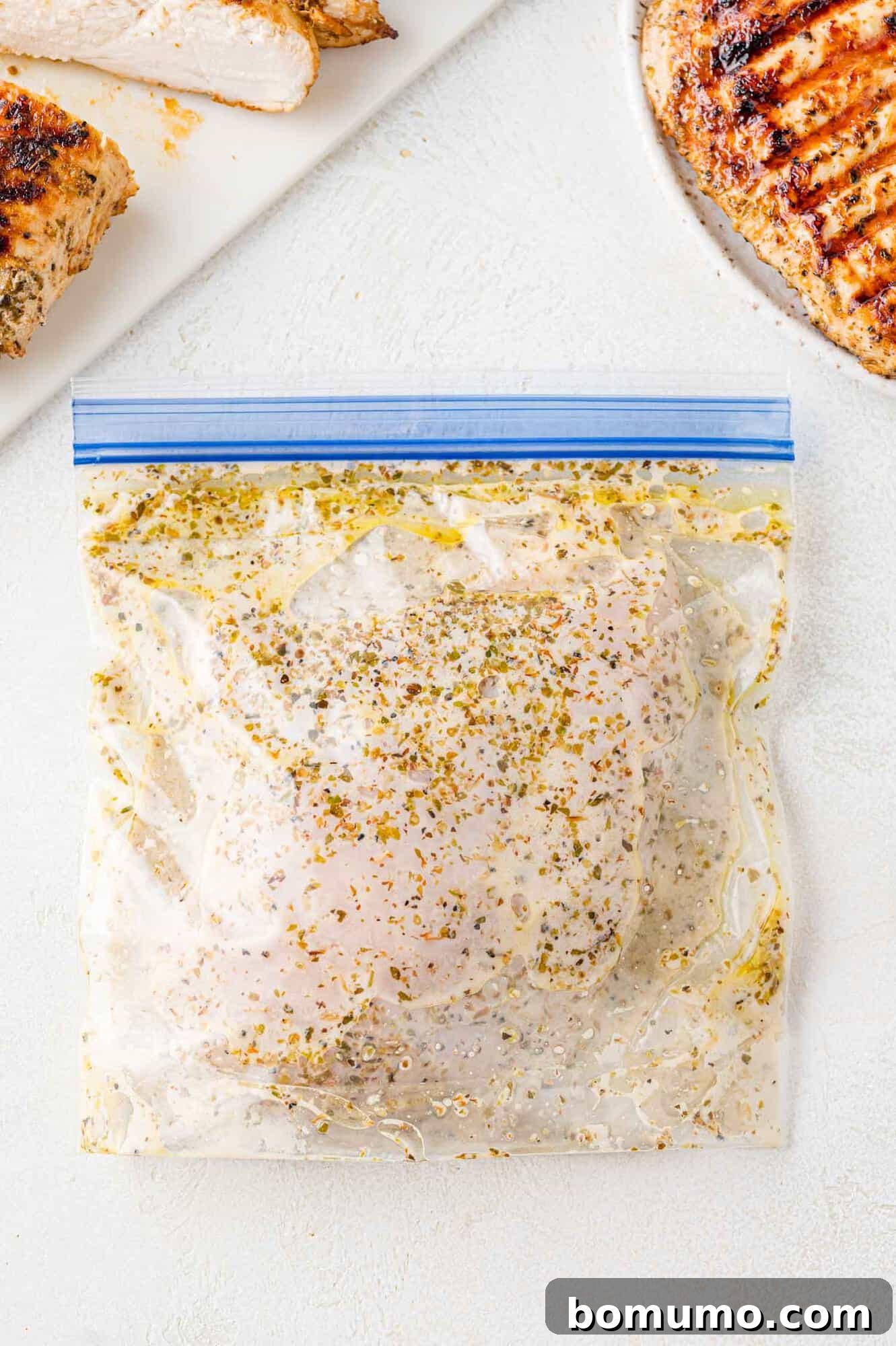 Chicken in Greek chicken marinade, in a zip-top bag, surrounded by grilled chicken, ready to be cooked or enjoyed.