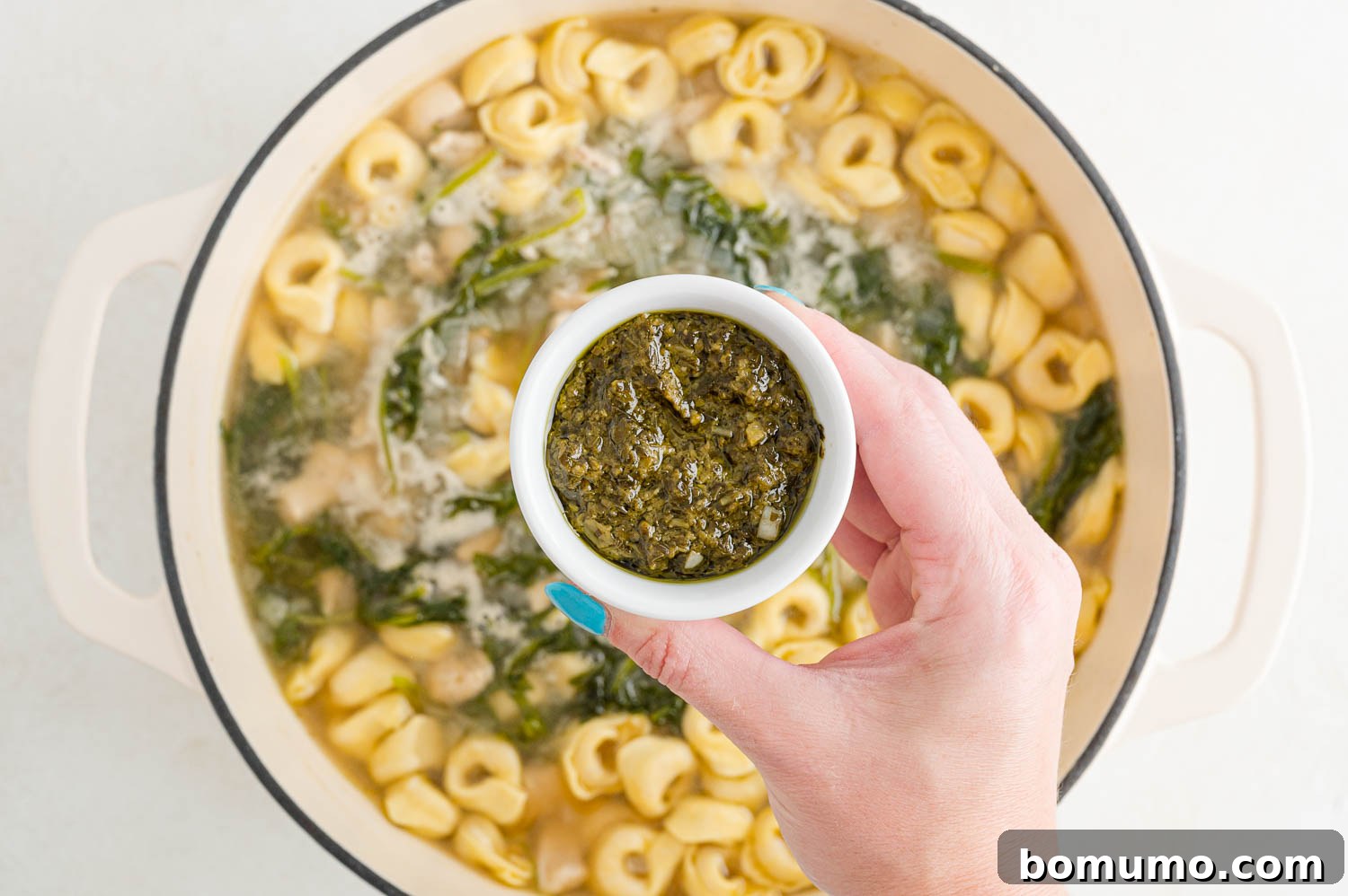 A dollop of vibrant green pesto being stirred into the hot chicken tortellini soup, adding a burst of flavor.