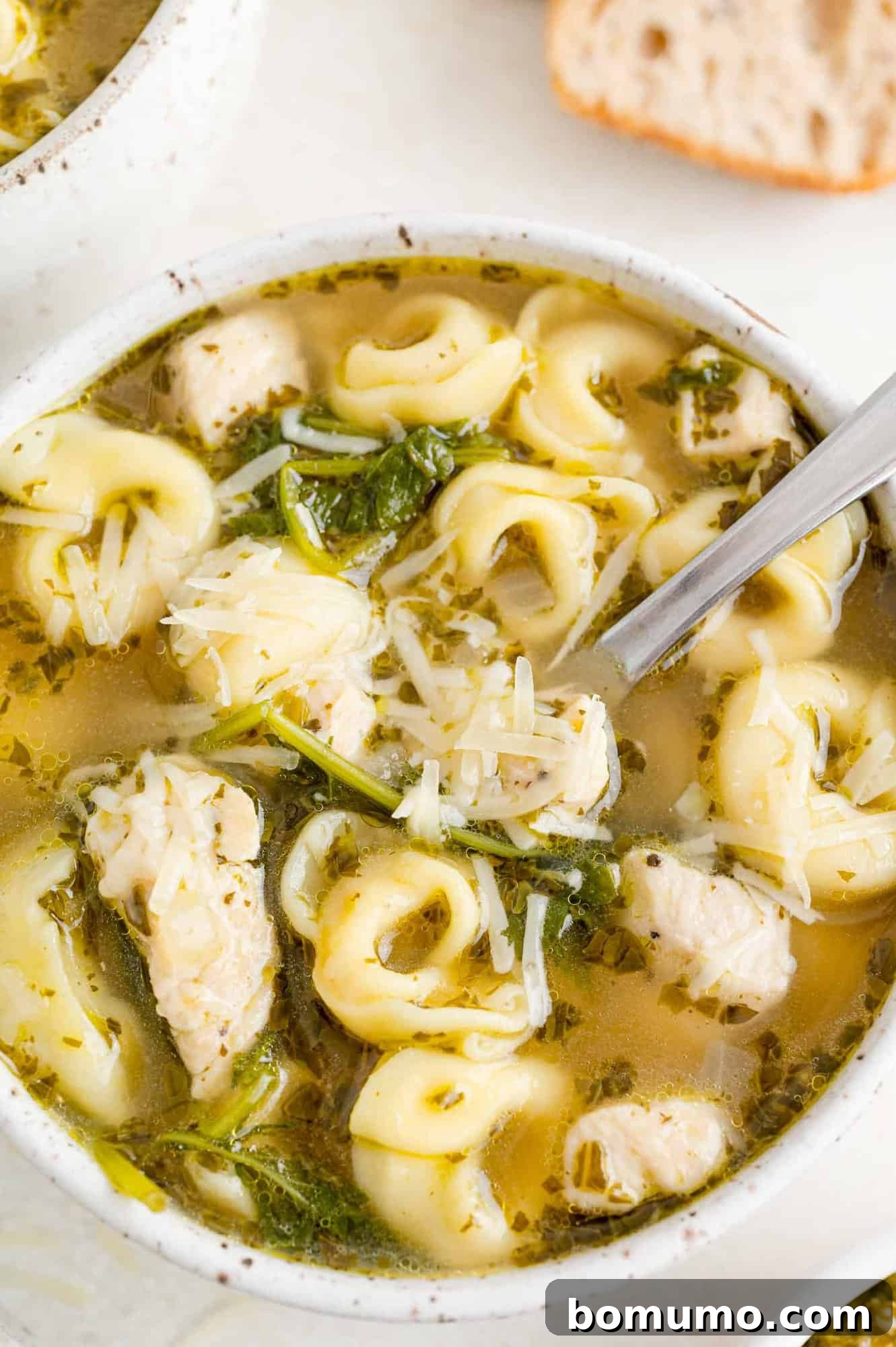 A warm bowl of chicken tortellini soup with kale and pesto, perfectly complemented by a slice of crusty bread, ready to be enjoyed.