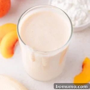 A glass of creamy peach cottage cheese smoothie sits on a white surface, with fresh peach slices and a bowl of cottage cheese beside it, ready to be enjoyed.
