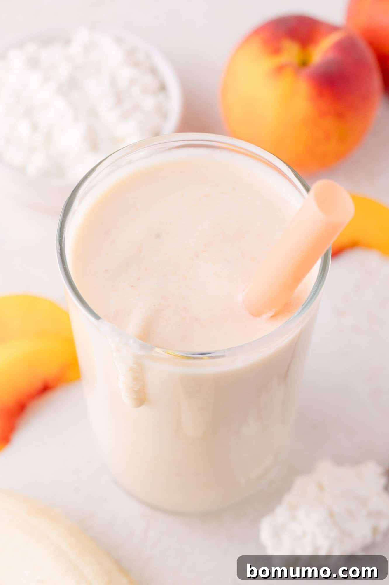 A beautifully composed shot featuring a glass of creamy smoothie with a straw, artfully arranged with fresh peach slices, a whole ripe peach, a bowl of cottage cheese, and a banana on a pristine white surface, highlighting the smoothie's fresh ingredients.