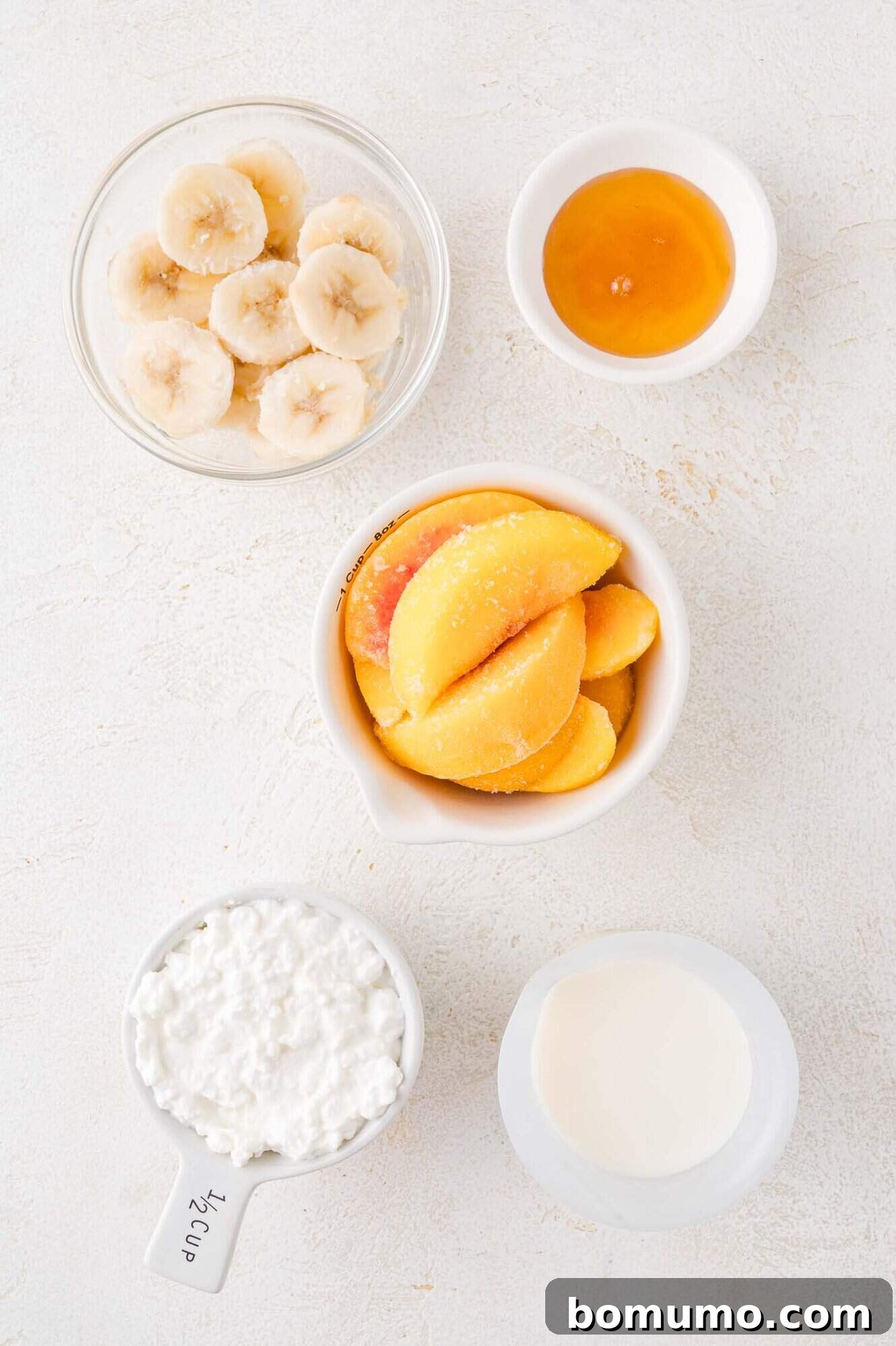 A top-down, artfully arranged view of all the smoothie ingredients: neatly sliced bananas, vibrant peach slices, a golden drizzle of honey, a creamy scoop of cottage cheese, and a splash of milk, each in its own small bowl on a crisp white surface, ready for blending.
