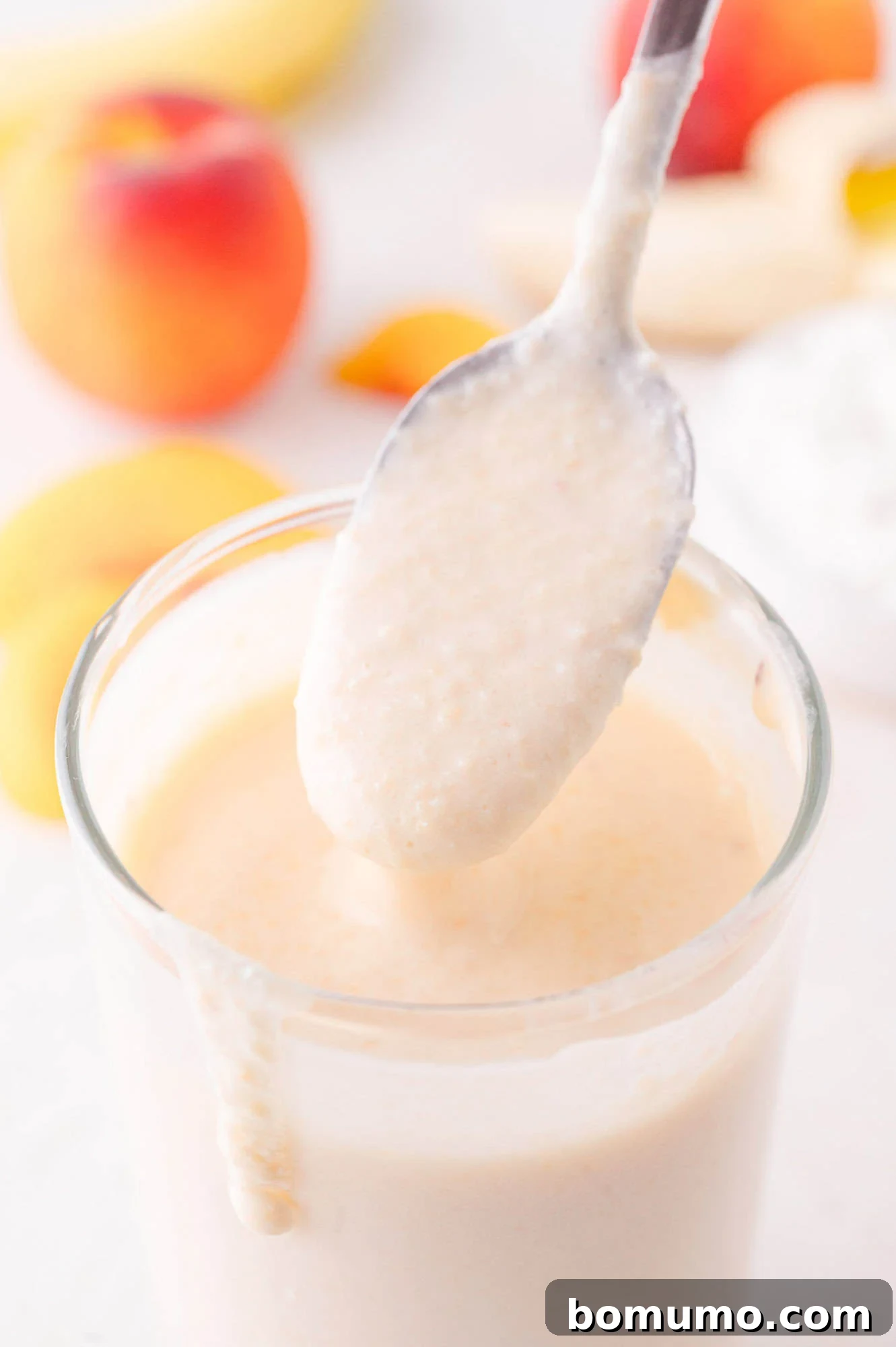 A spoon elegantly lifts a thick, creamy spoonful of smoothie above a glass, showcasing its rich texture, with blurred peaches, bananas, and peach slices in the background creating a fresh and inviting scene.