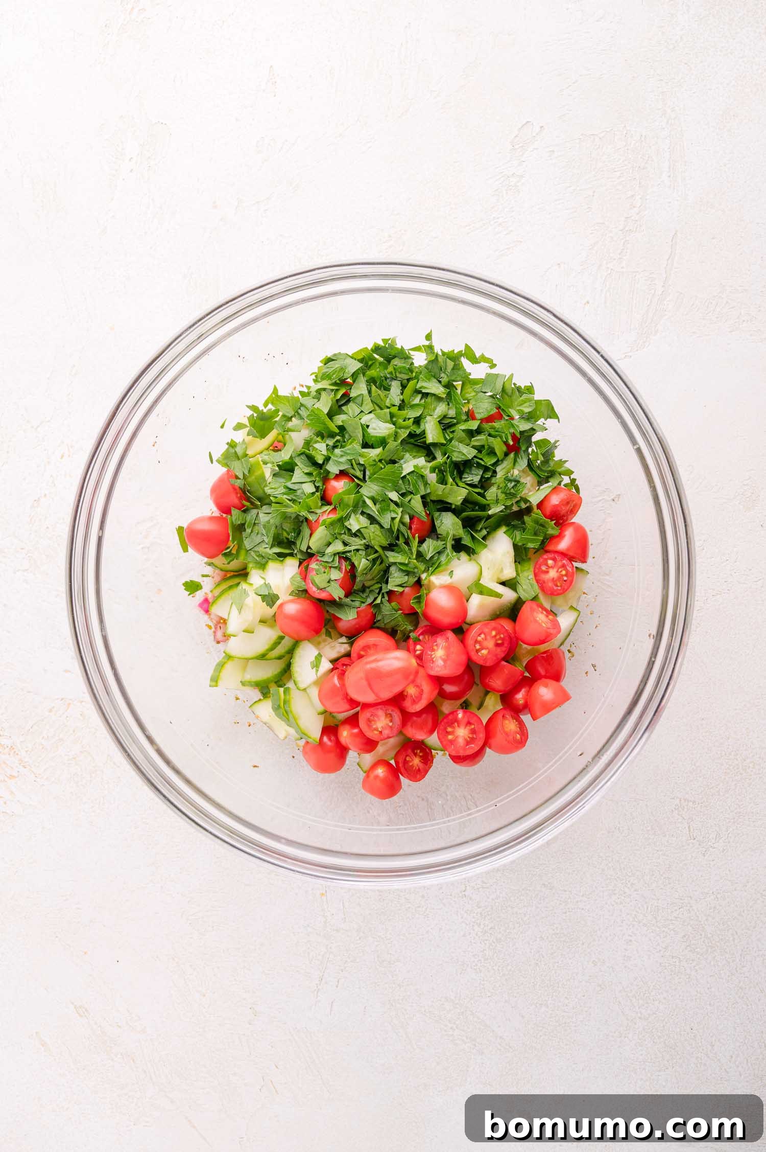 A large clear glass bowl filled with a fresh mixture of chopped tomatoes, crisp cucumbers, and vibrant parsley, glistening with vinaigrette on a pristine white surface.